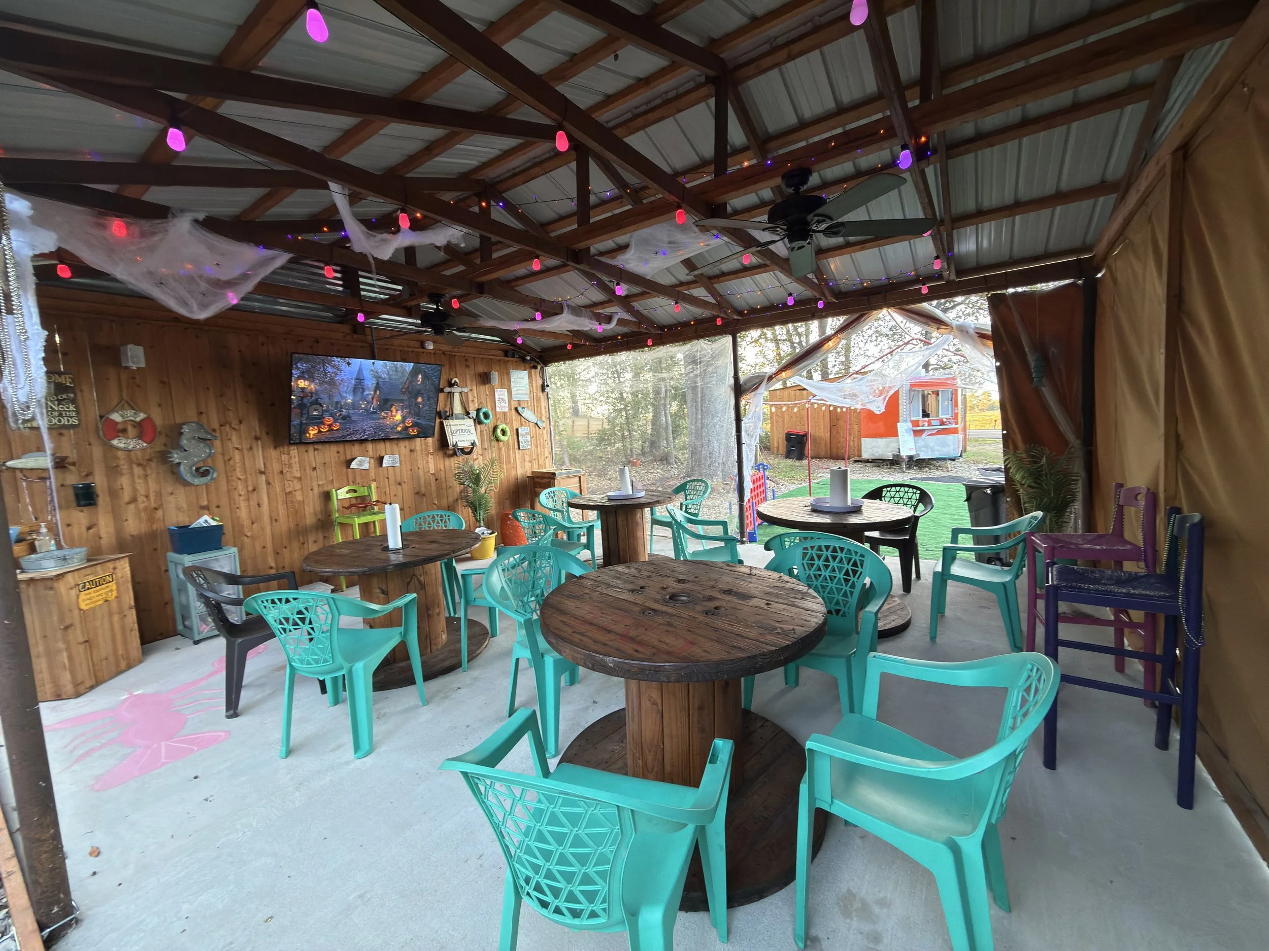 Decorated outdoor party area with colorful chairs, spider web decorations, and a large TV mounted on the wooden wall, with a food truck outside.