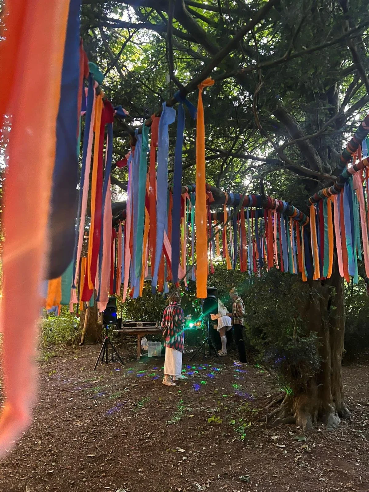 People gathering in a wooded outdoor space with a large tree draped in colorful fabric strips. Two individuals are near a small DJ setup with speakers, one is wearing a white dress and the other a patterned jacket. Green laser lights are projected onto the ground and surrounding area.