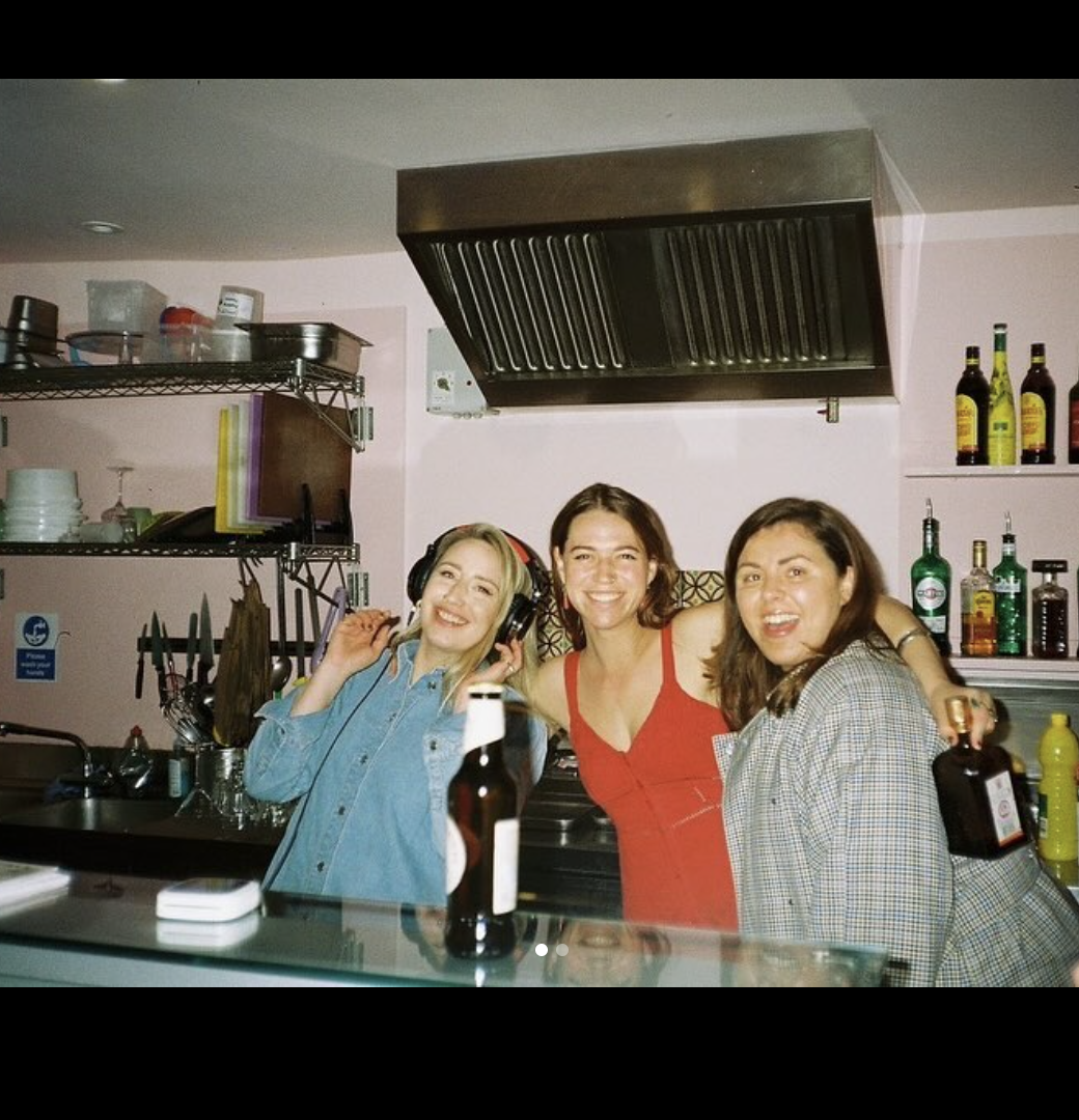 Three women smiling and enjoying themselves behind a bar counter with bottles and bar tools, in a kitchen or bar setting.