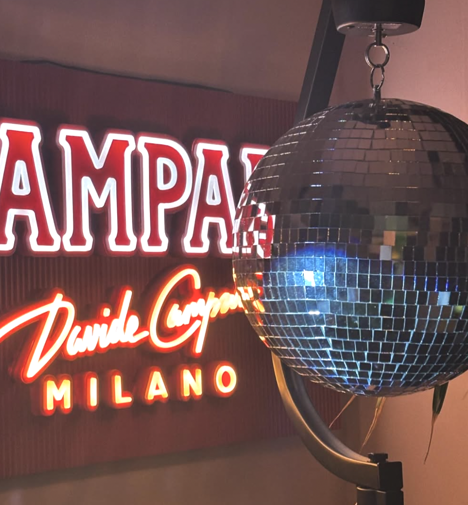 A mirrored disco ball hanging in front of a neon sign that reads 'TAMPA' and 'Dame Cucina MILANO' in red and white neon lights.