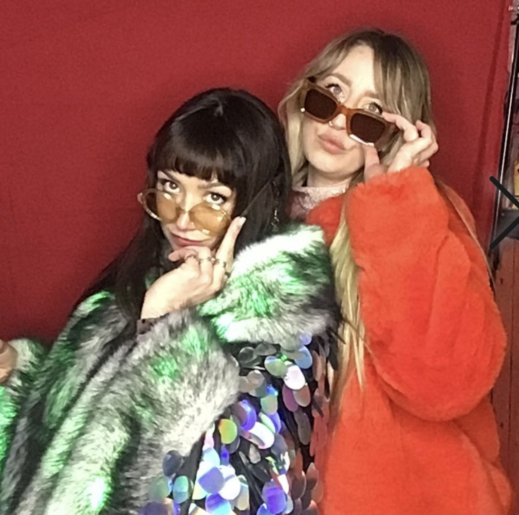 Two women posing in front of a red background, both wearing sunglasses and colorful furry coats.