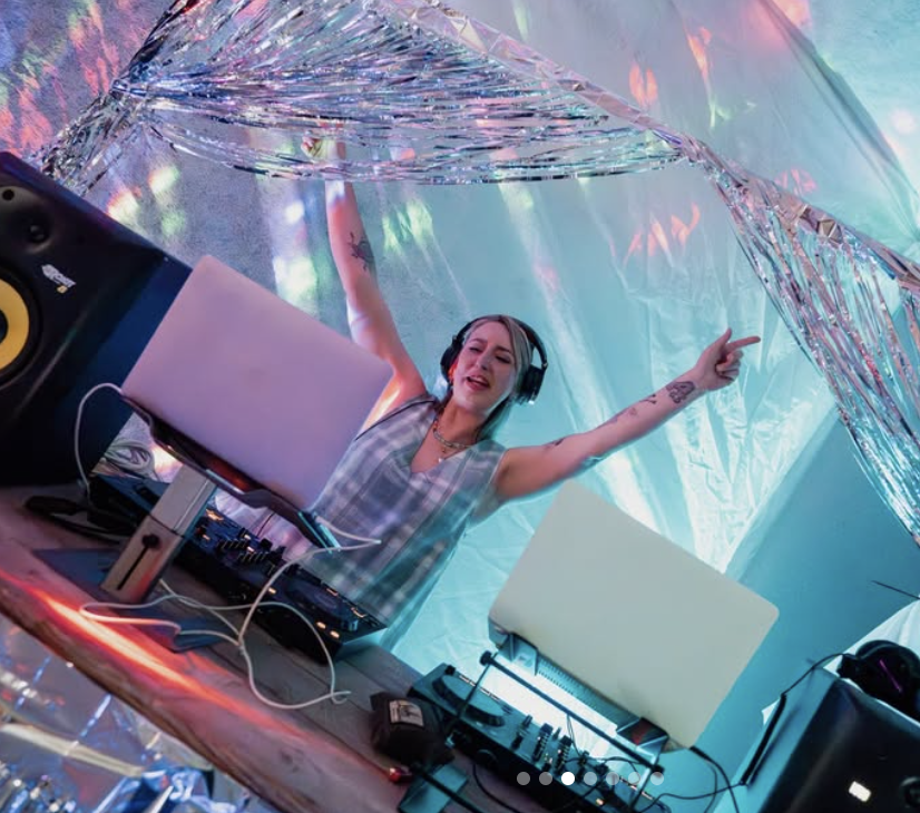Alice DJ with headphones, smiling and dancing, at a colourful, iridescent party setup with DJ equipment and laptops.