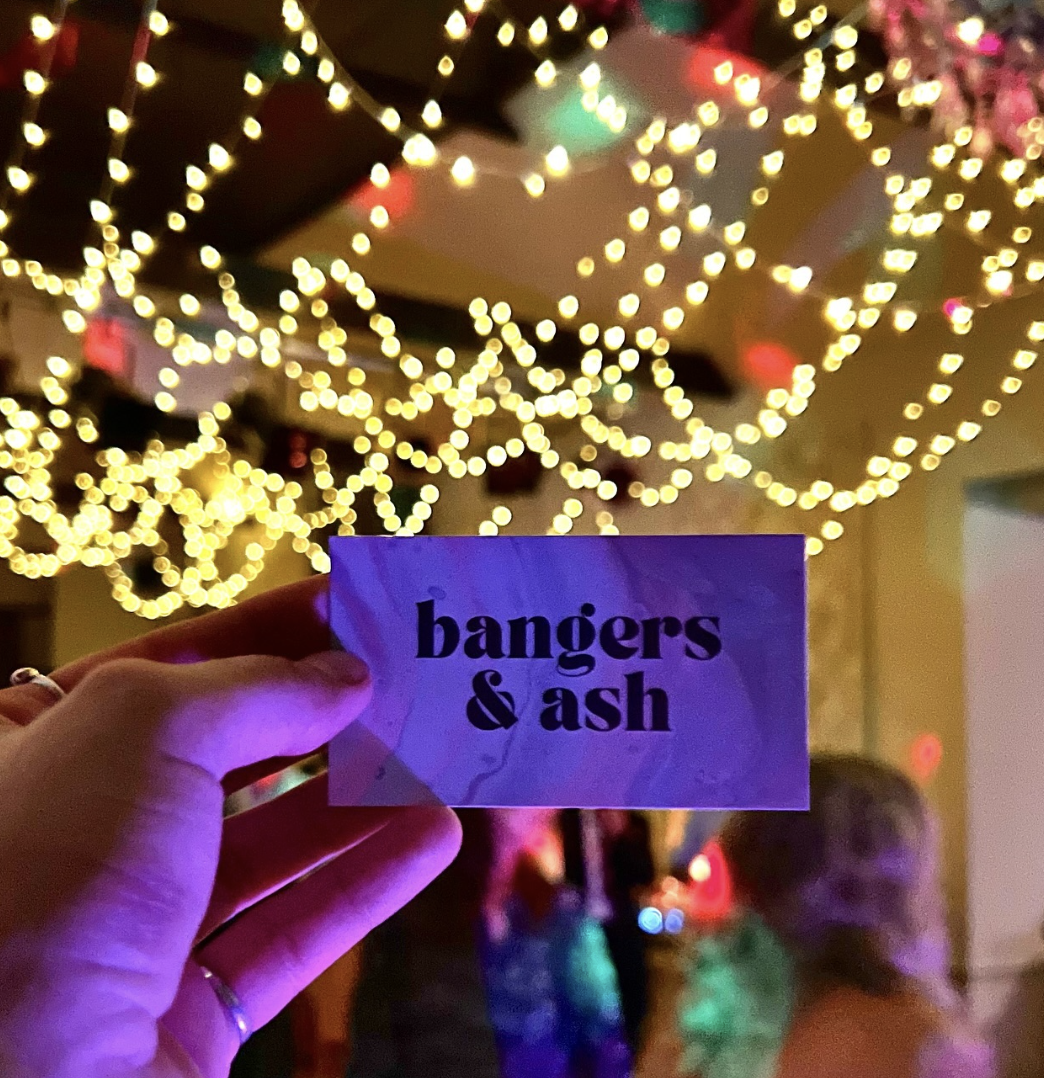 A hand holding a small card with the words 'bangers & ash' in front of a background of warm-colored string lights and balloons, suggesting a festive or party setting.