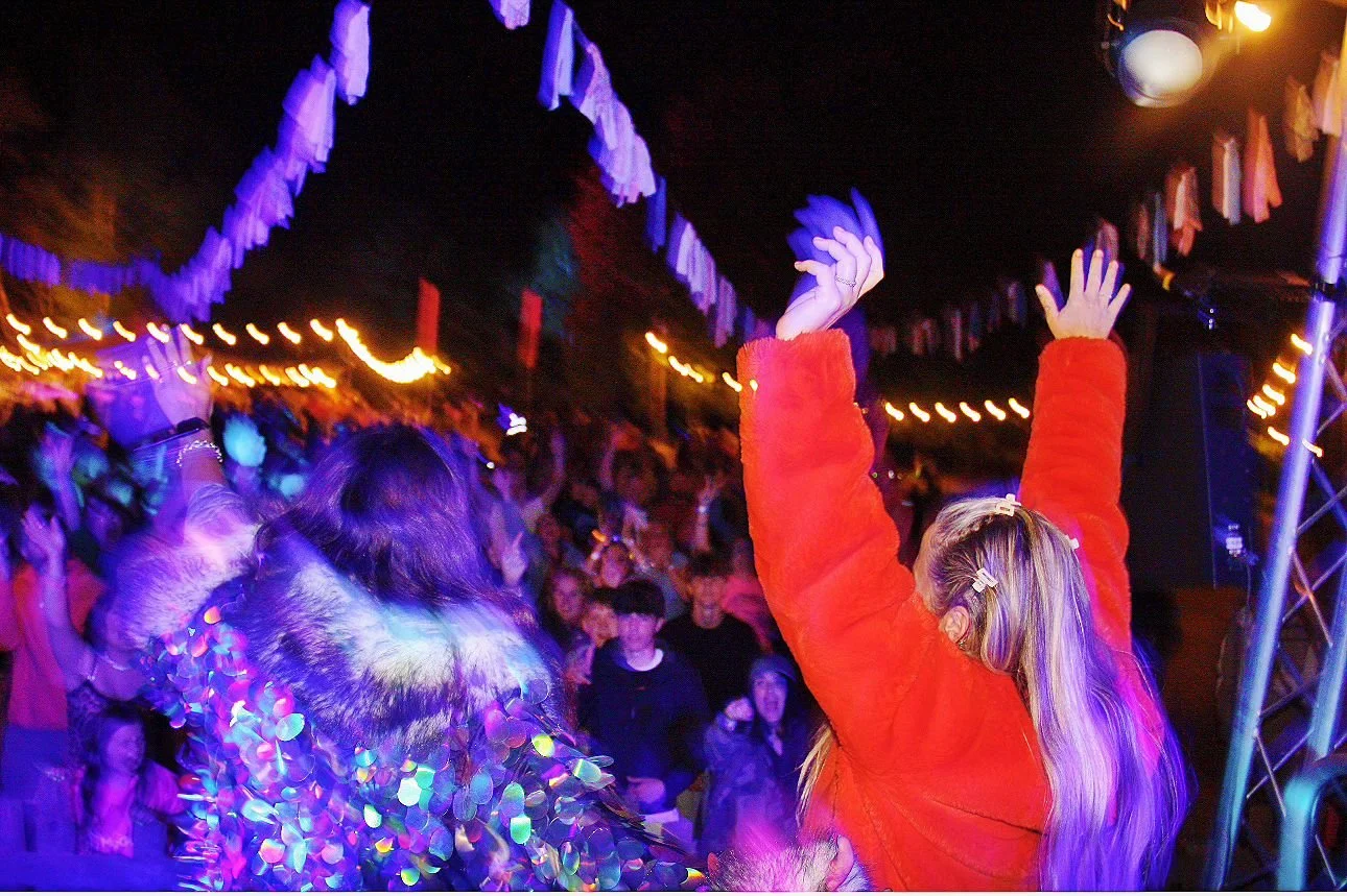 People dancing at a party or concert with colorful lights, night setting, and decorative hanging banners.
