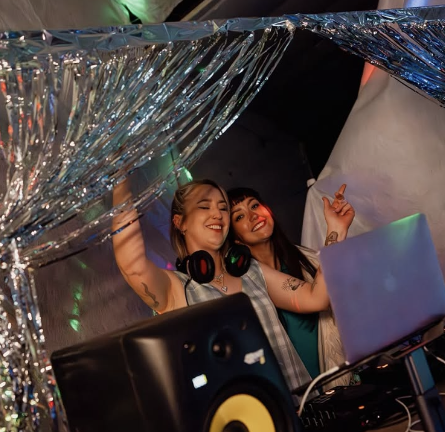 Two women DJing at a party, surrounded by shiny silver decorations, with colorful lights, a laptop, and DJ equipment.