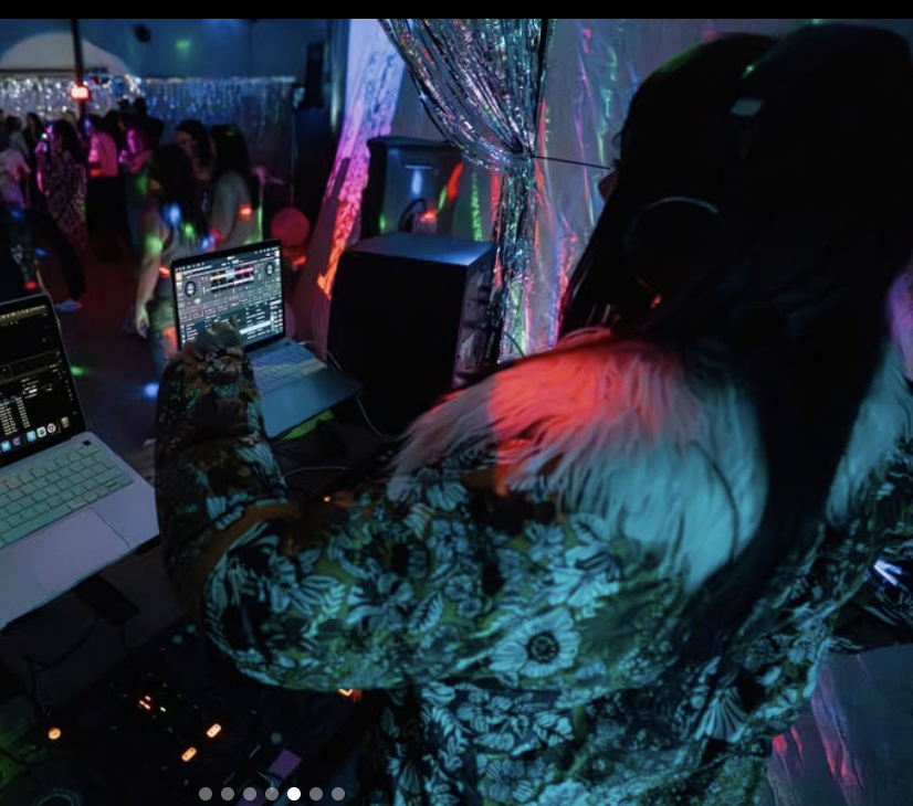 DJ performing at a nightclub with equipment including laptops and speakers, colorful lights, and a crowd dancing in the background.