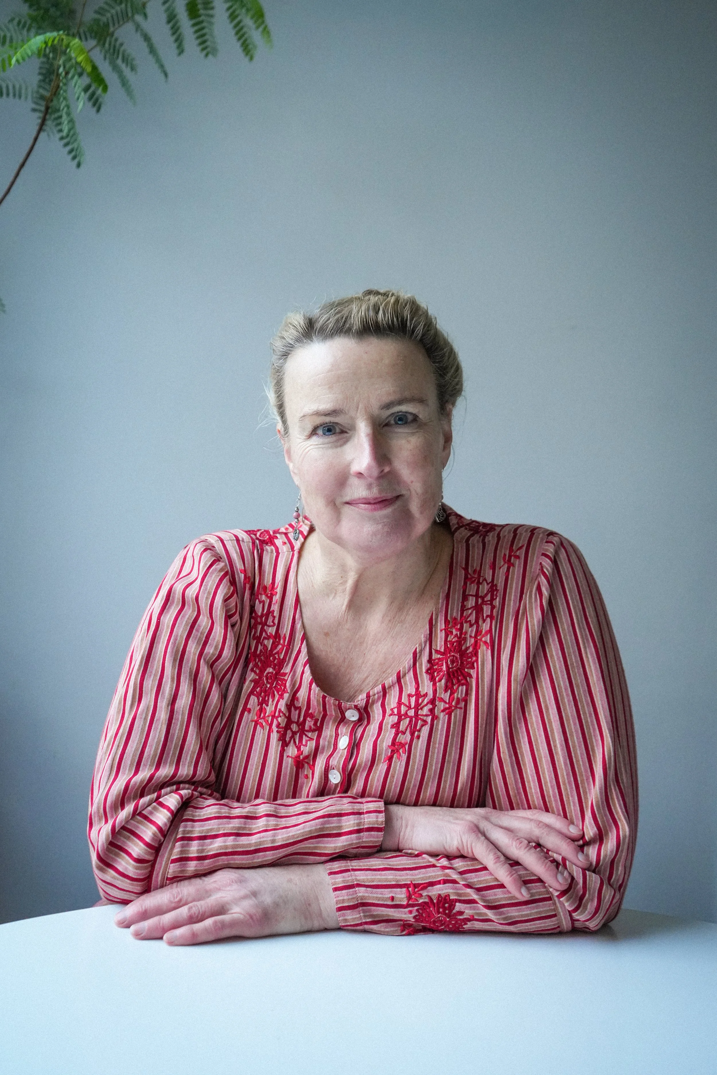 A middle-aged woman sitting at a white table with her arms crossed, wearing a red and pink striped blouse with embroidered floral details, in front of a plain gray background with green leaves in the upper left corner.