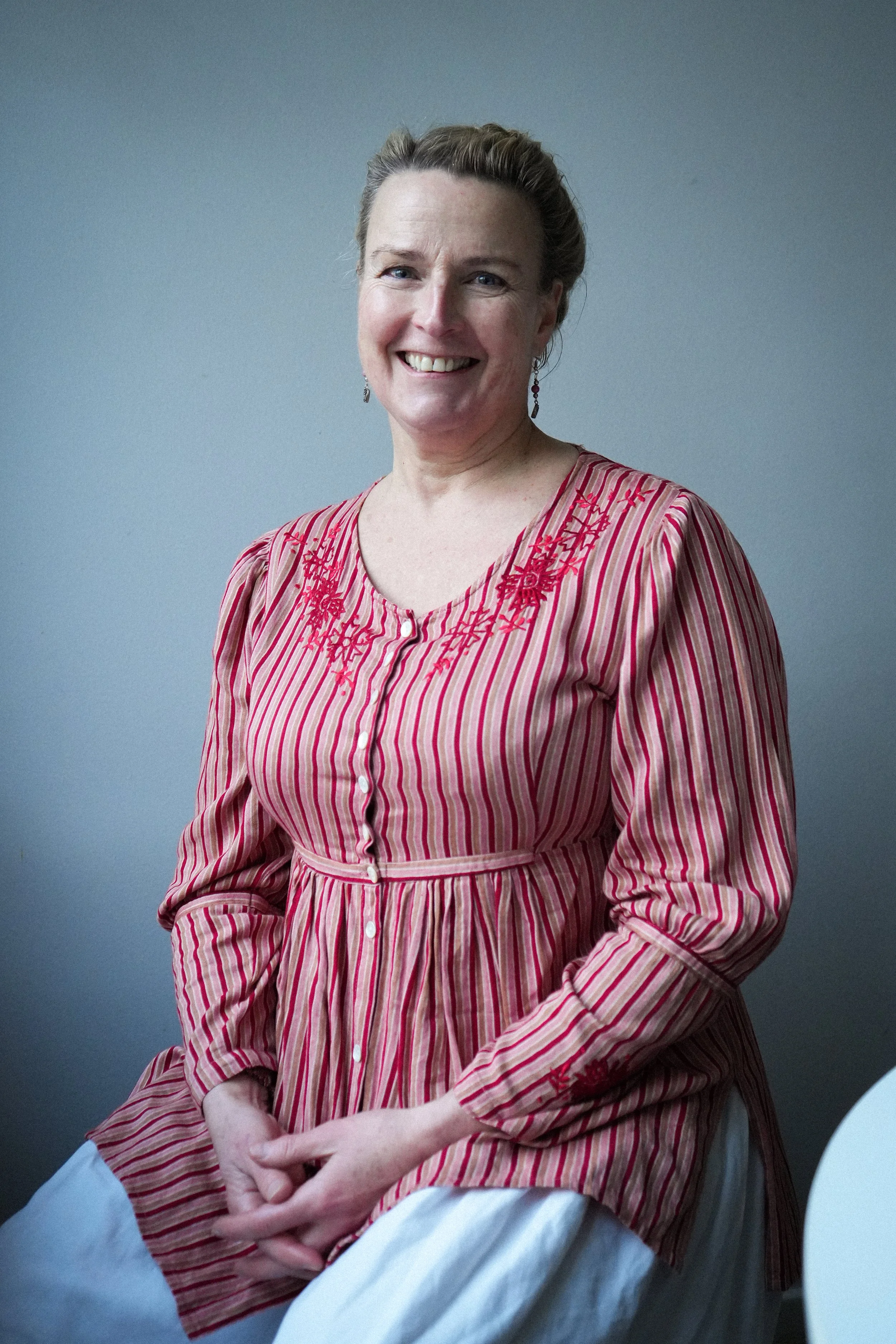 A woman with blonde hair in an updo, smiling, wearing earrings and a red and white striped dress with embroidered details, sitting against a plain background.