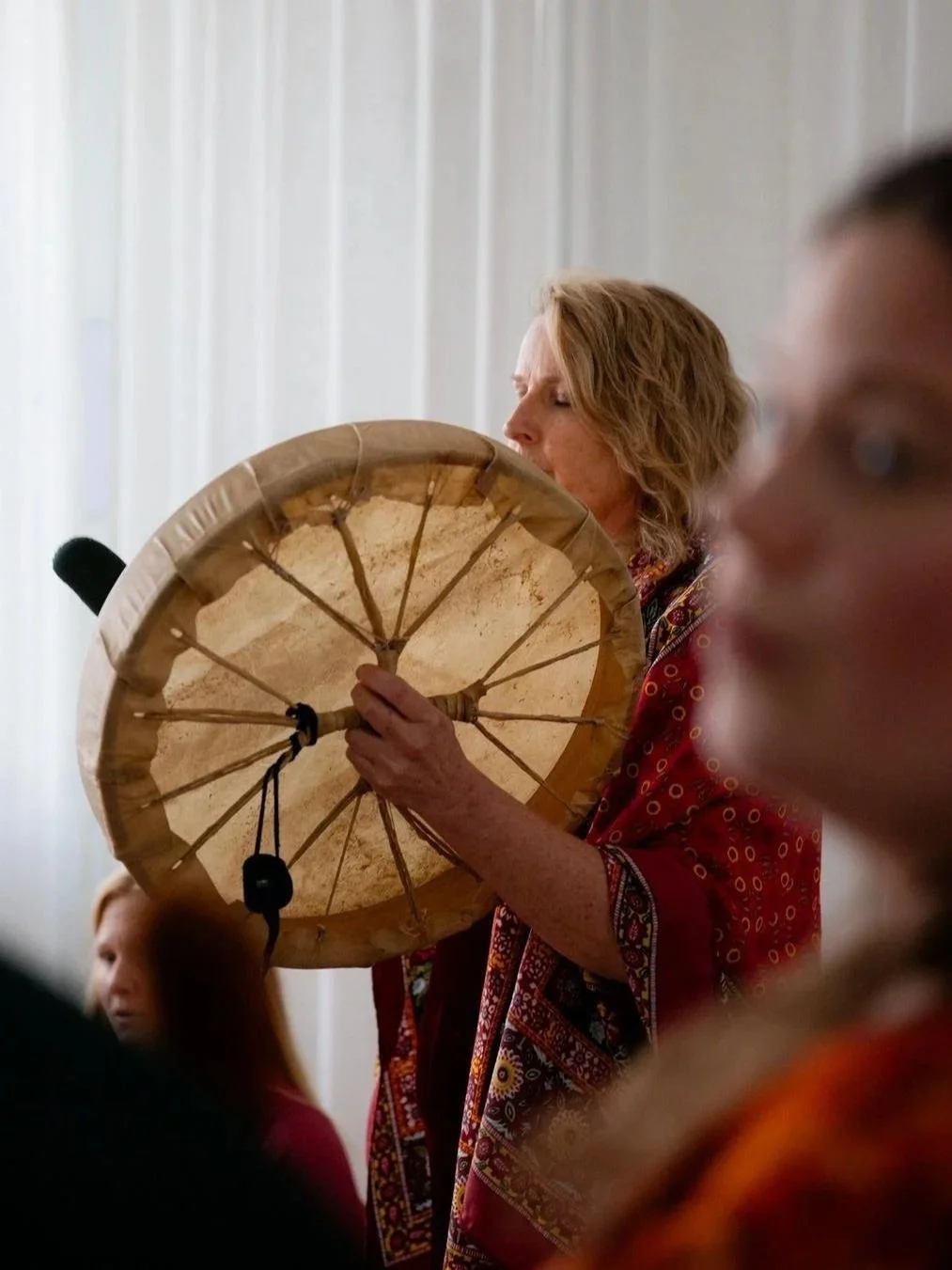 A woman with curly blonde hair in a red patterned shirt appears to be playing a large drum or tambourine, which she is holding up to her face. She is in an indoor setting with white vertical blinds in the background. Several other people are partially visible in the foreground.