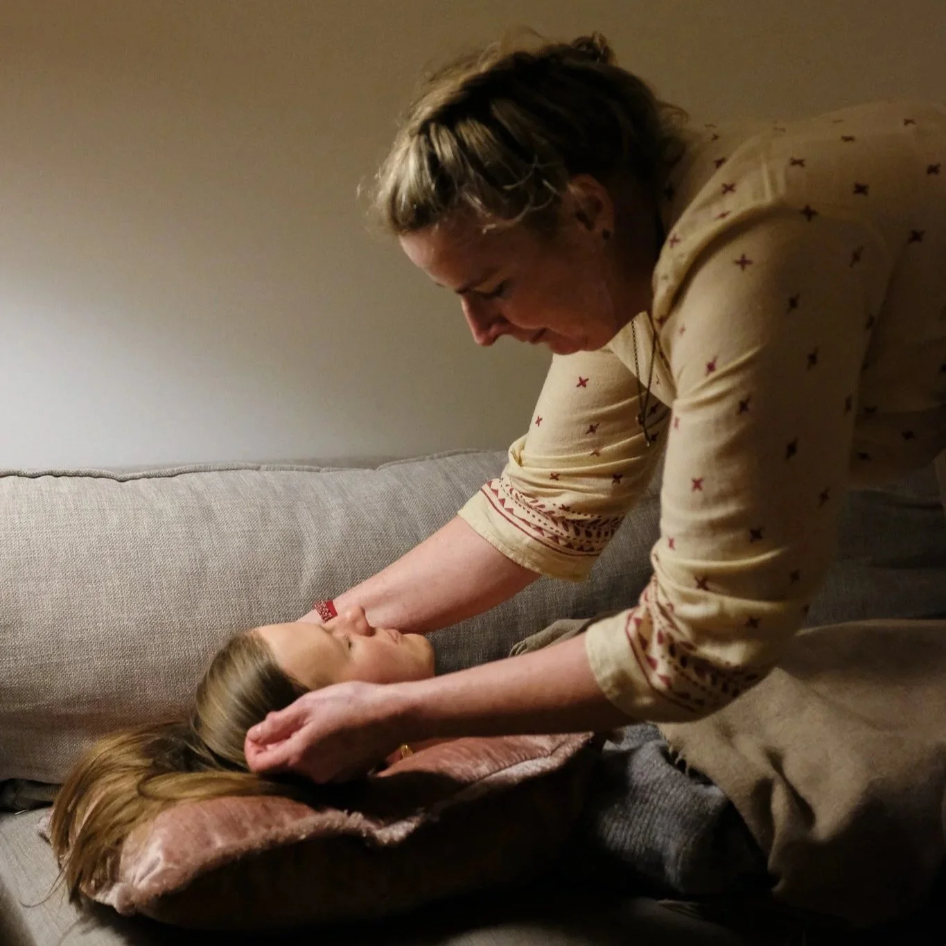 A woman leaning over a couch, gently touching the face of a sleeping young girl lying on a pillow, with both appearing to be in a comforting moment.