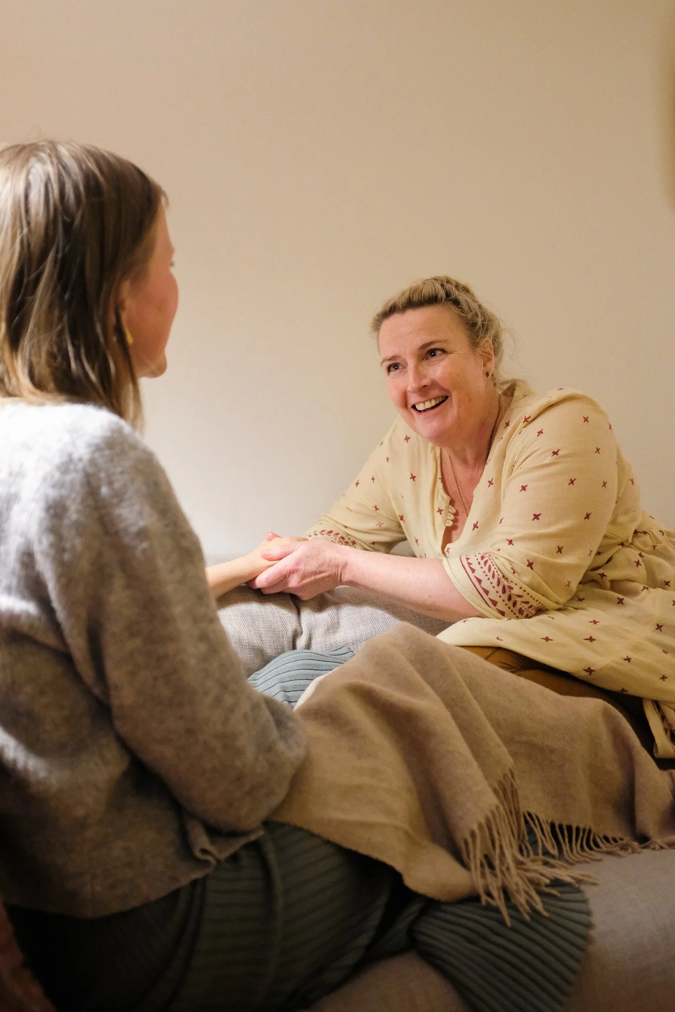 Two women having a friendly conversation, one is smiling and the other is partially turned away from the camera, sitting indoors against a plain wall.