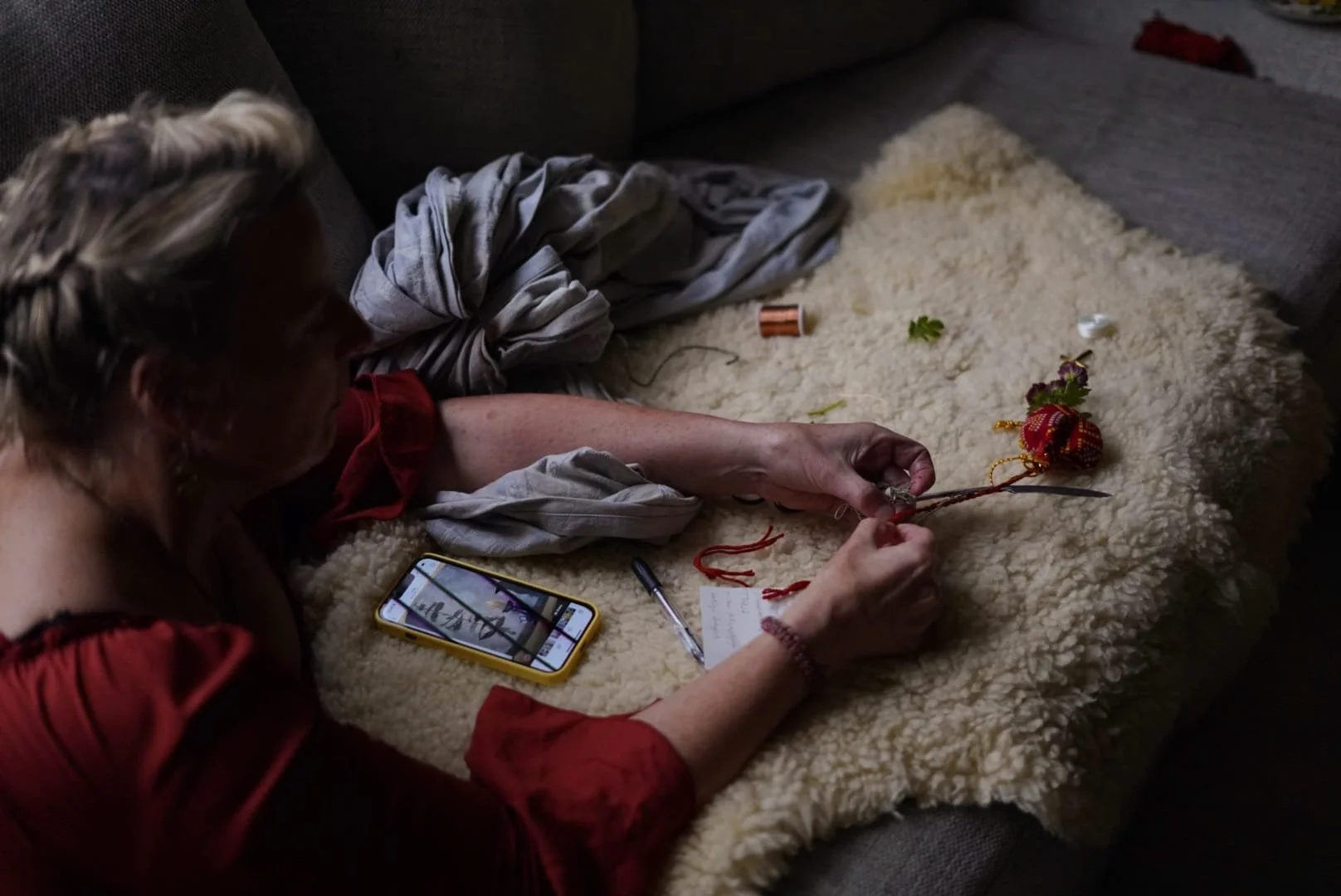 A person laying on a couch with a fluffy beige blanket, holding Christmas ornaments and decorations, with a smartphone nearby on the blanket and a writing utensil.
