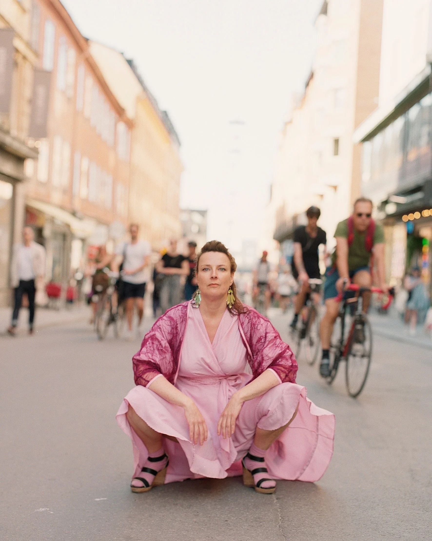 A woman dressed in pink and purple traditional dress squats on the ground with her hands resting on her knees. She is on a city street with blurred pedestrians and buildings in the background.