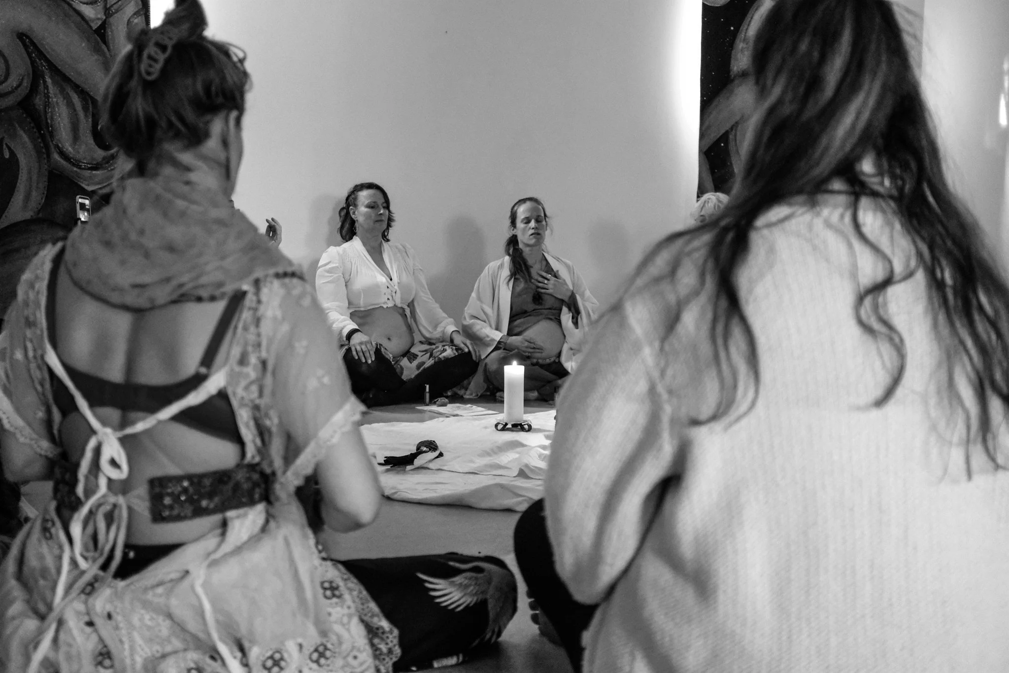 A group of women participating in a meditation or prayer session, sitting on the floor with a lit candle in front of them. Two women in the center are pregnant, with one placing her hand on her belly and the other holding her hand on her chest, suggesting a spiritual or reflective gathering.
