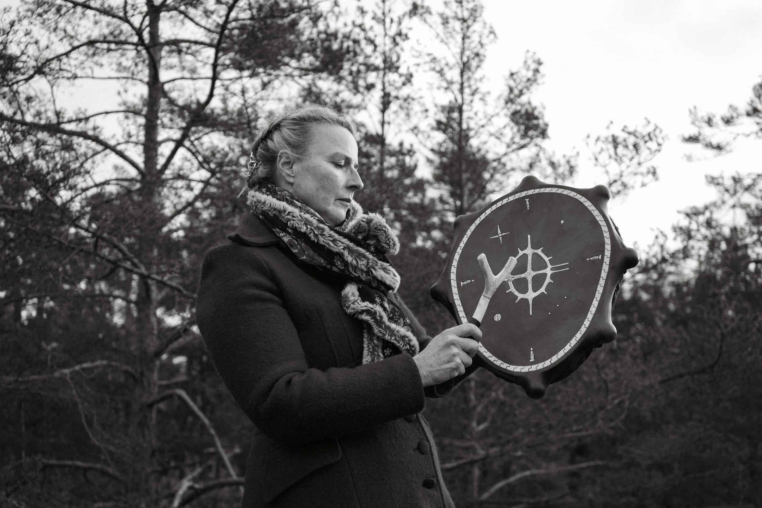 A woman with braided hair and a floral scarf appears to be holding or examining a decorative shield with a mystical or geometric design on it, outdoors among trees.