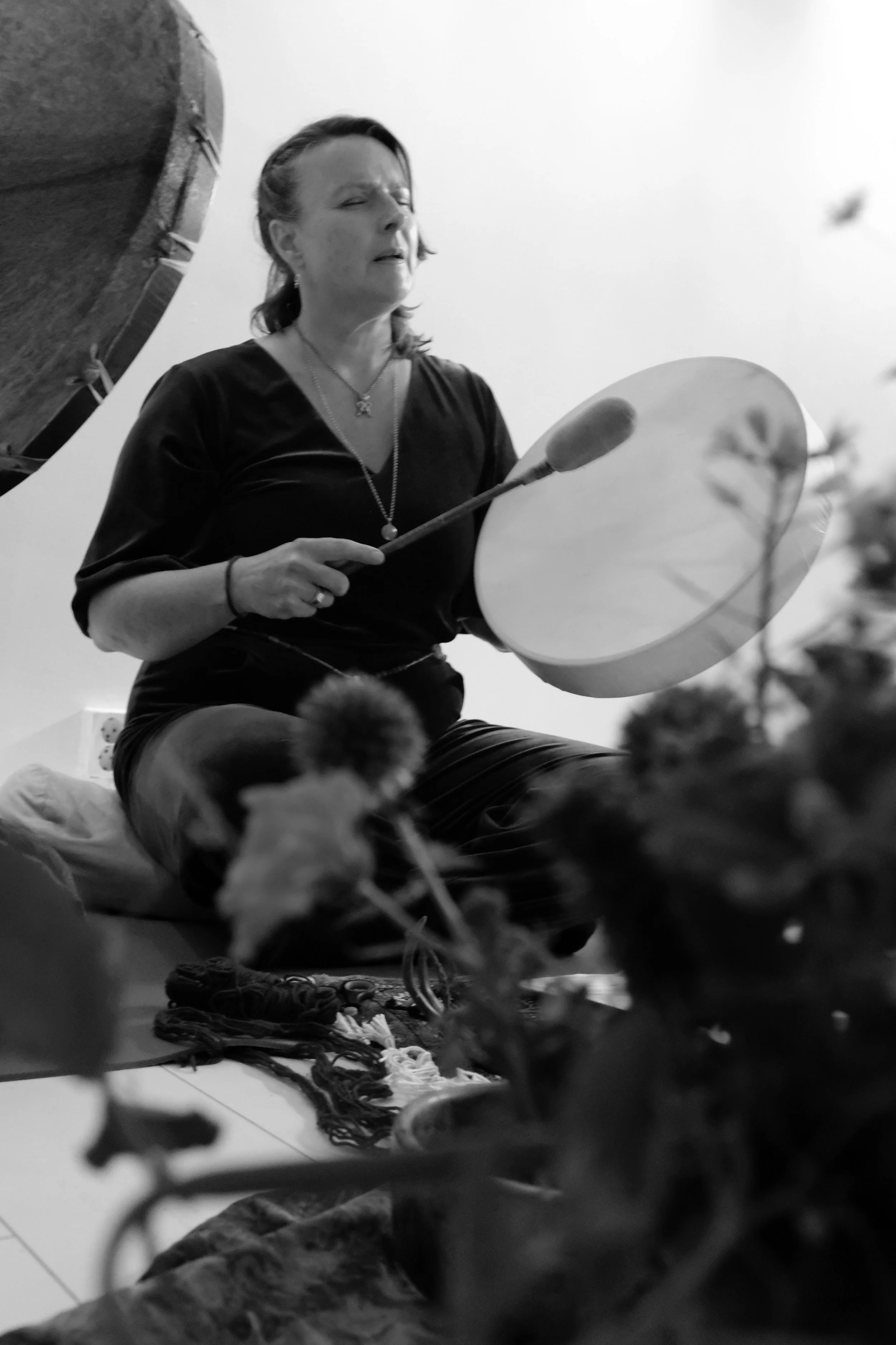 A woman in a black dress sitting on the floor, playing a percussion instrument with a drumstick, with yarn and a pair of shoes nearby, in an indoor setting.