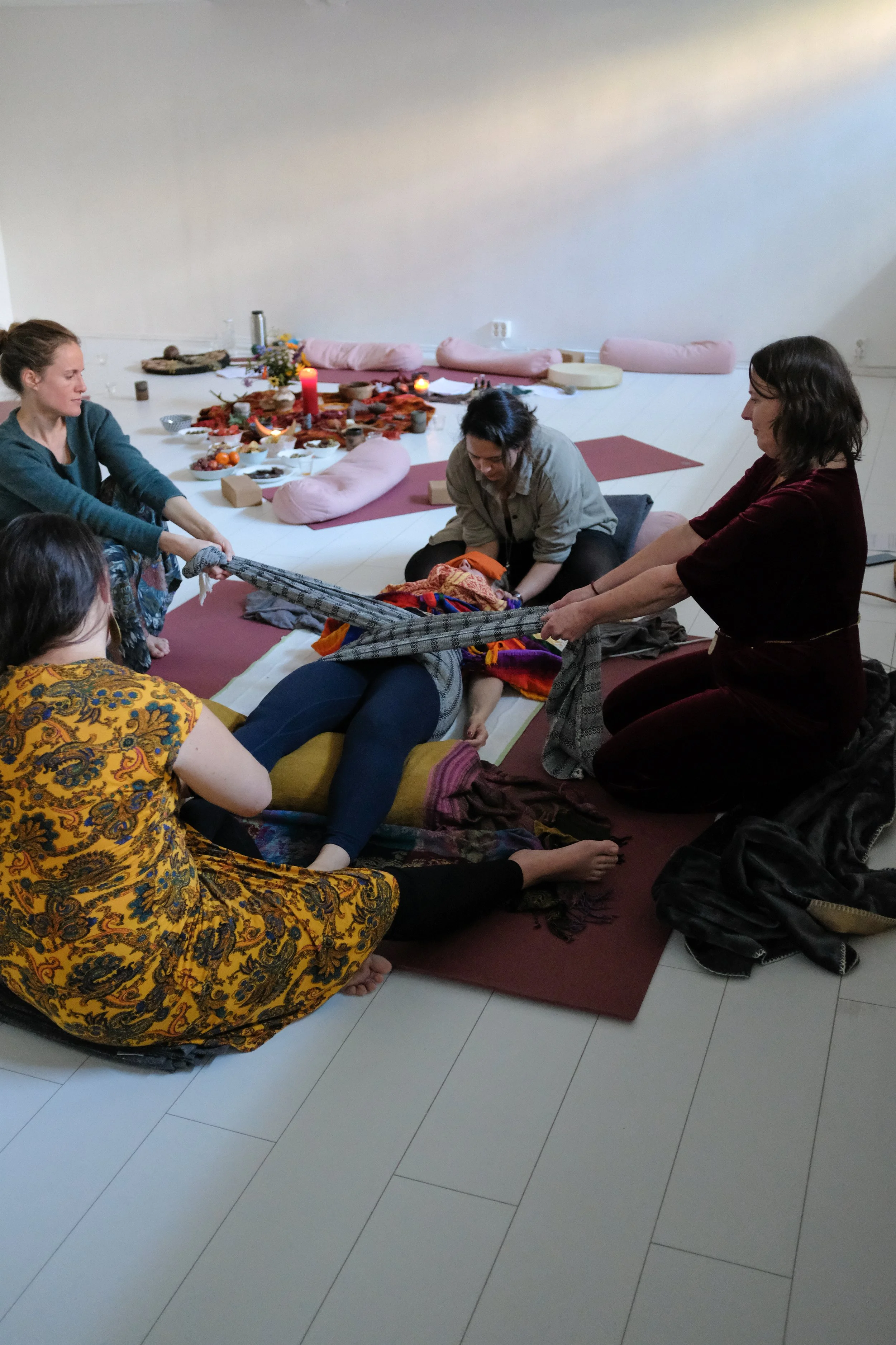 A group of people sitting on the floor participating in a spiritual or meditation session. The setting includes cushions, a low table with offerings, candles, and flowers, creating a serene atmosphere.
