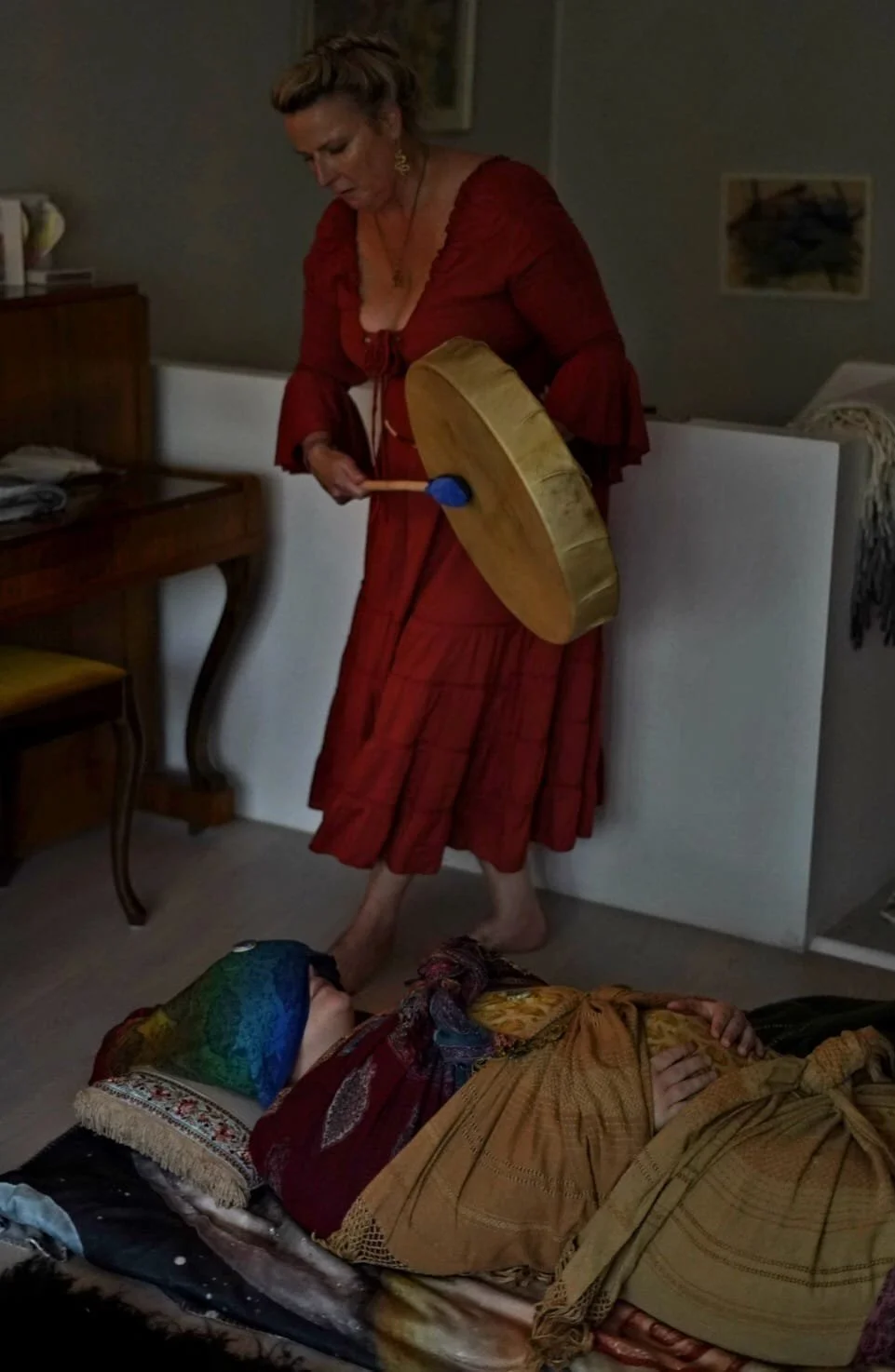 A woman in a red dress is standing, holding a drumstick, and looking down at a person lying on the floor dressed in colorful clothing with a headscarf. The person on the floor appears to be unconscious or asleep. The setting is an indoor room with furniture and artwork on the walls.