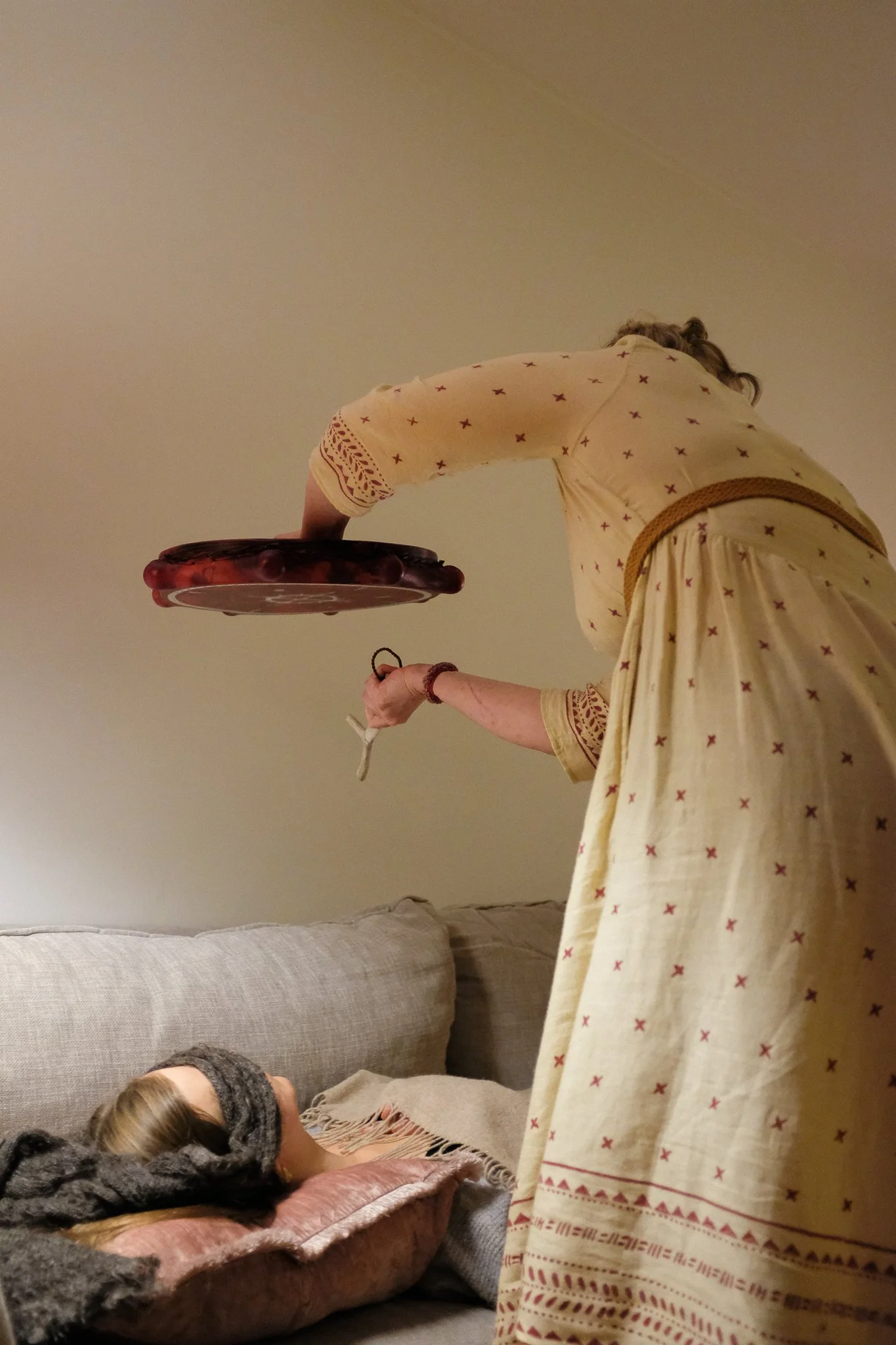 A woman in a cream-colored dress with red star patterns and lace details on the sleeves and hem, standing and reaching towards a cake on a table, with a person lying on a couch in front of her.