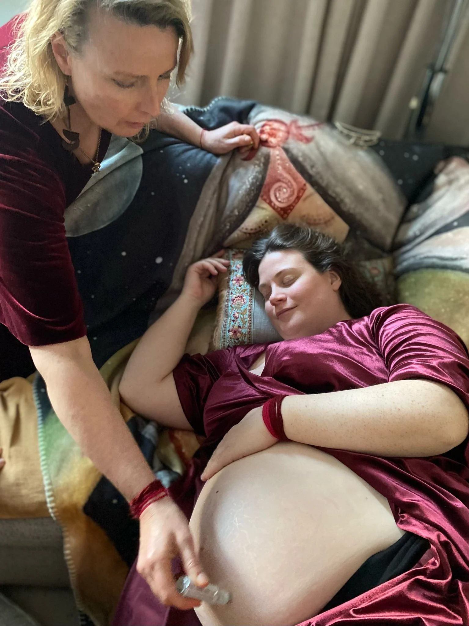 A woman in a velvet pink top lying down with her eyes closed, receiving a massage or treatment on her thigh from another woman in a dark top. They are indoors, with curtains in the background.