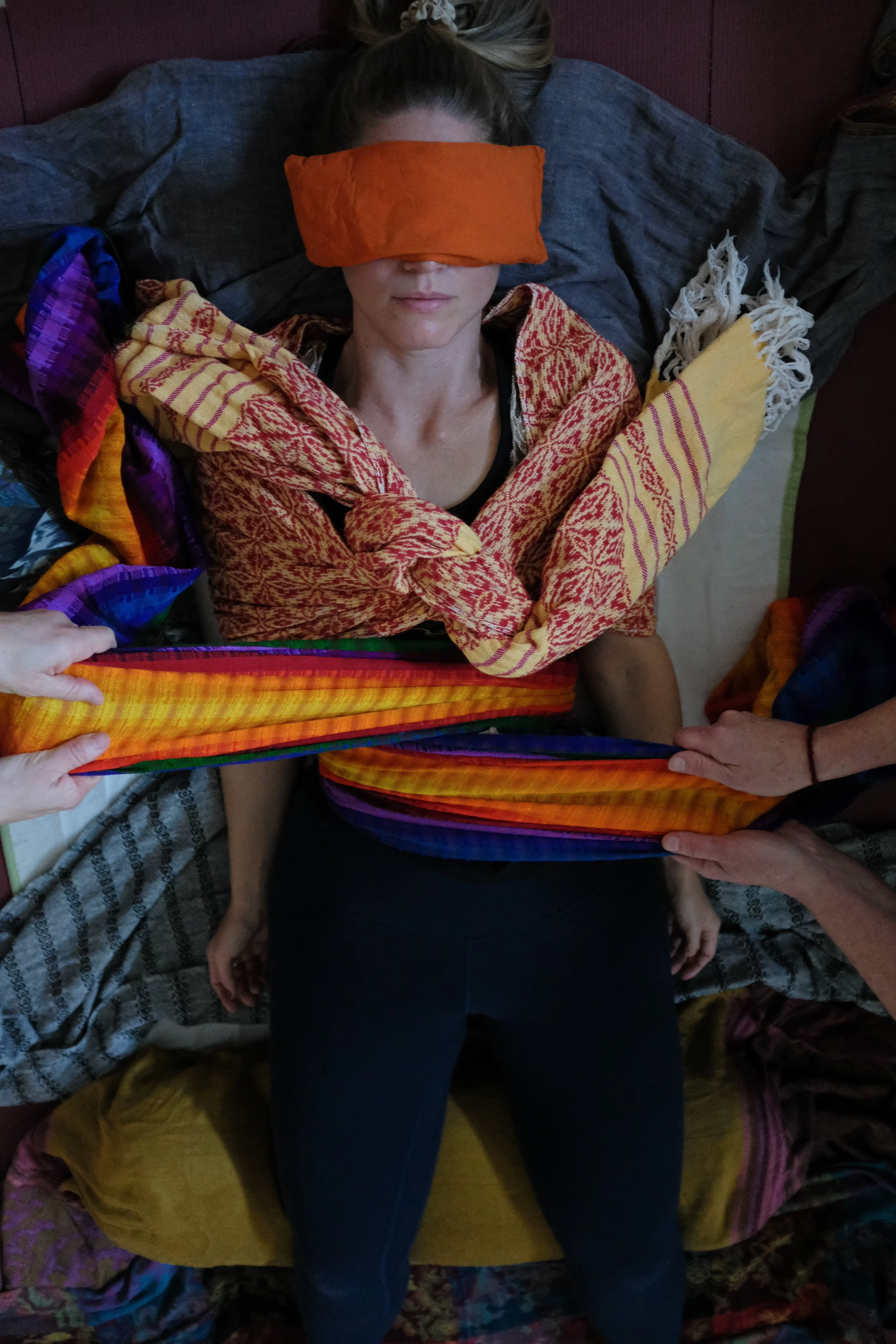 A person lies on a bed with colorful blankets, wearing a dark shirt and dark pants. Their head is covered with an orange cloth, and their eyes are closed. Multiple hands are holding a rainbow-striped fabric around the person.