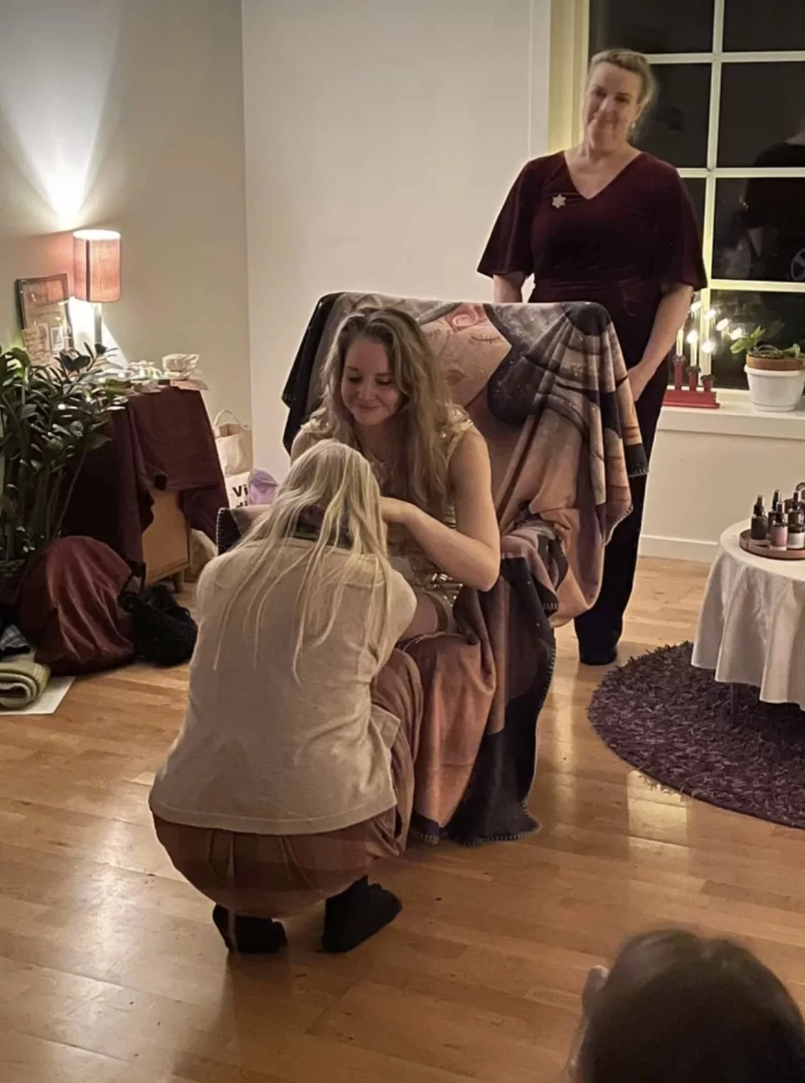 Three women are in a cozy, warmly lit room with wooden floors, one is sitting on a chair, another is crouching, and the third is standing nearby watching them. There are various decorative items and plants on a table and windowsill.