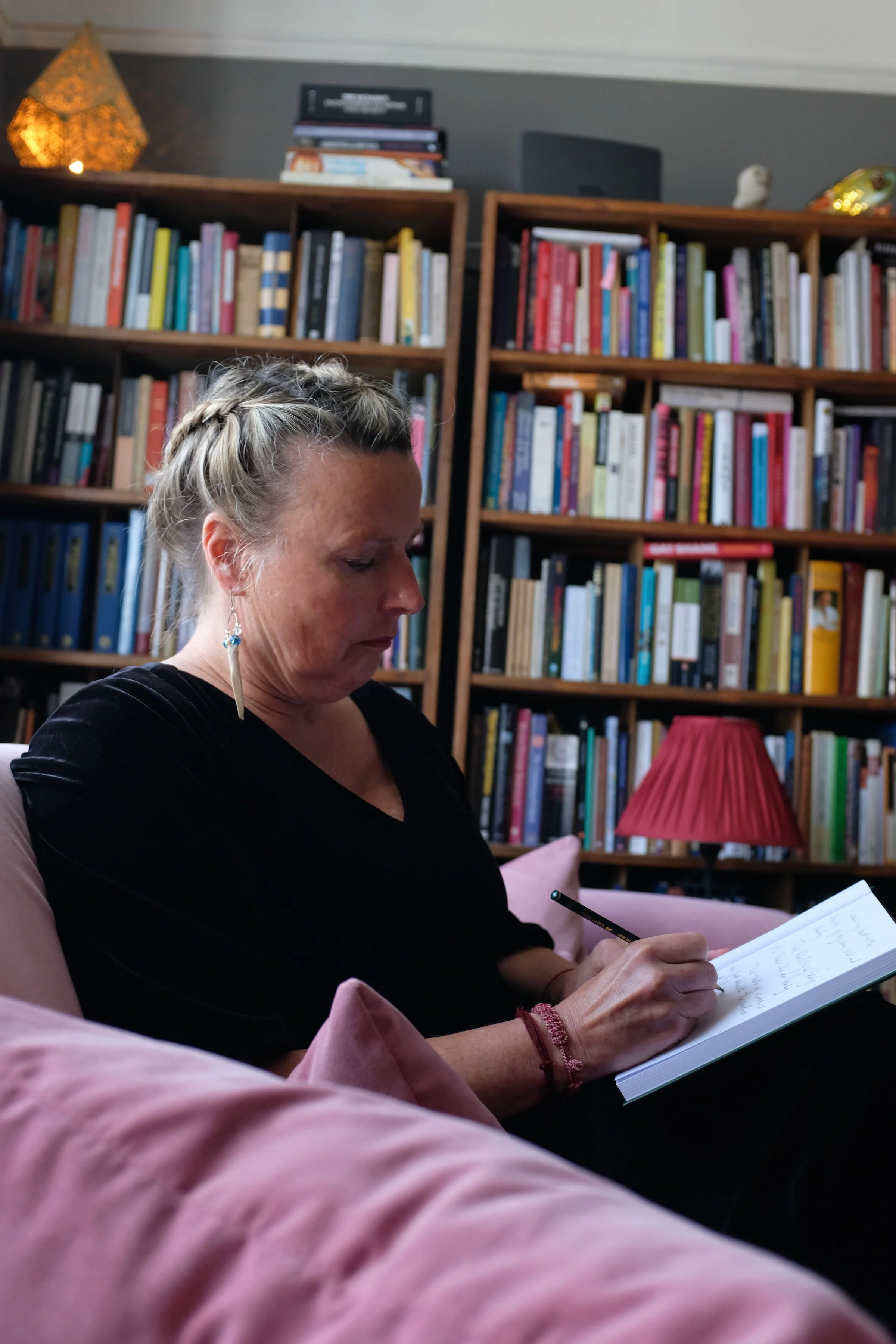 A woman with braided hair and earrings, sitting on a pink chair in front of a large bookshelf filled with books, writing in a notebook with a black pen, with a red lampshade in the foreground.