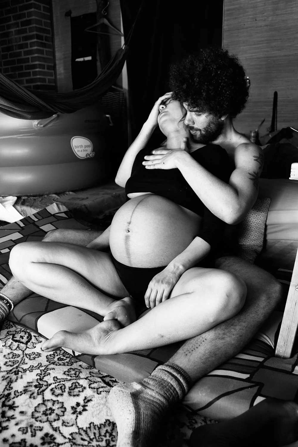 A couple sits closely together on a striped mat, embracing each other, with the woman visibly pregnant. The man holds the woman's head and gazes at her, while she rests her hand on his face. In the background, there is an inflatable birth pool and other household items.