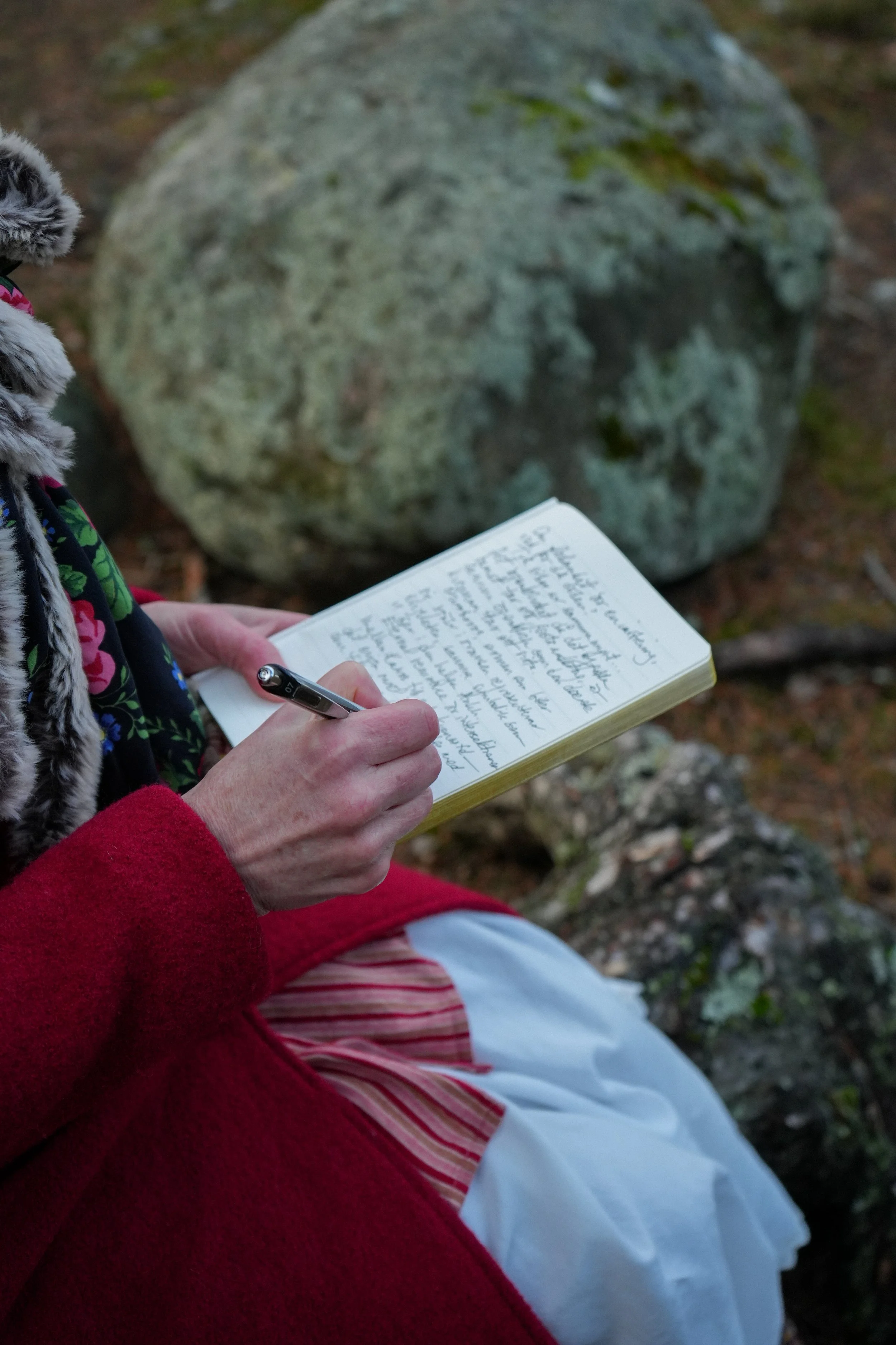 Person sitting outdoors on the ground, writing in a small notebook with a pen, large moss-covered rock in the background, surrounded by forest floor with leaves and moss.