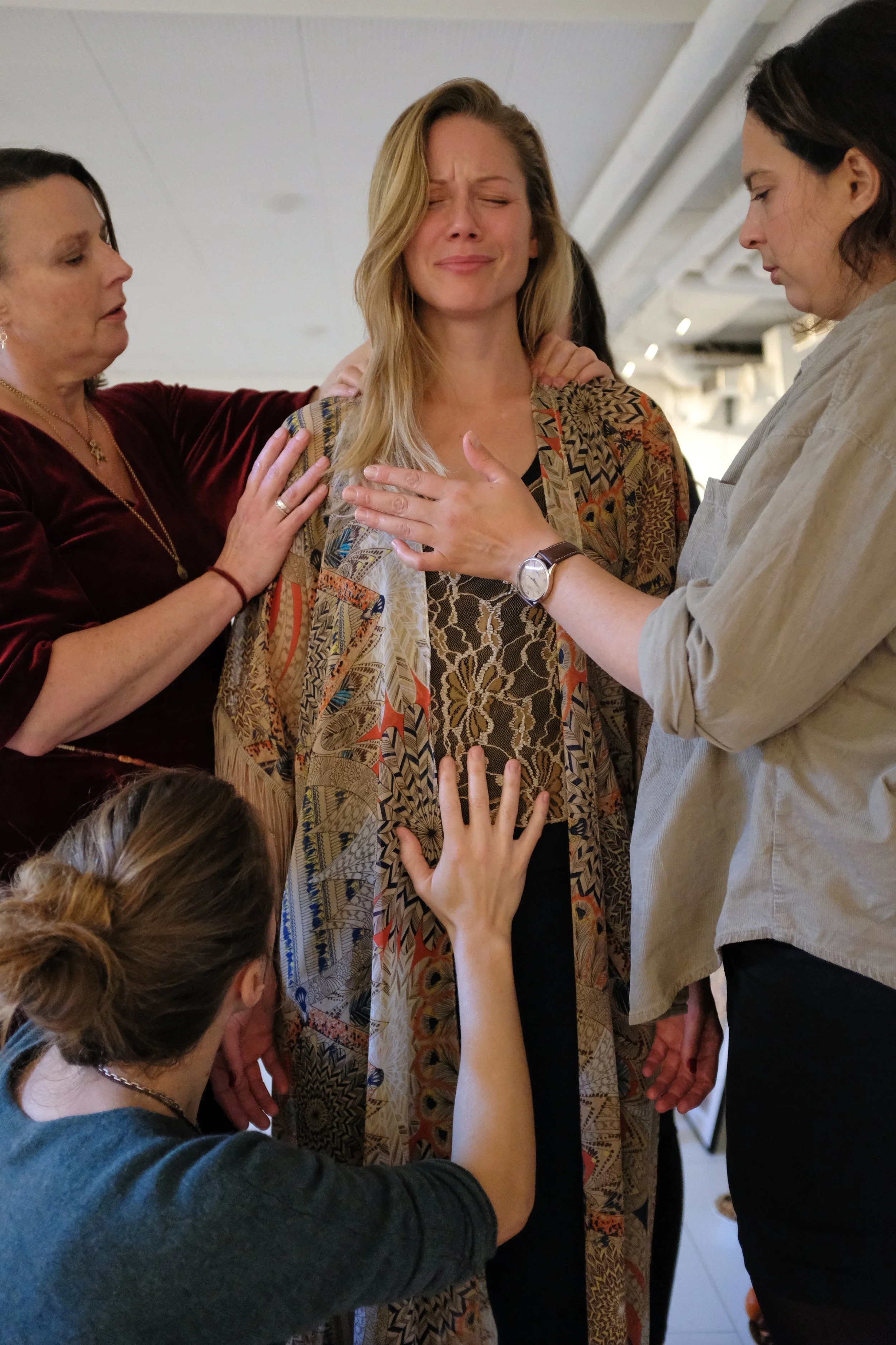 A woman standing with eyes closed and hand over her chest, surrounded by other women and children in an indoor setting.