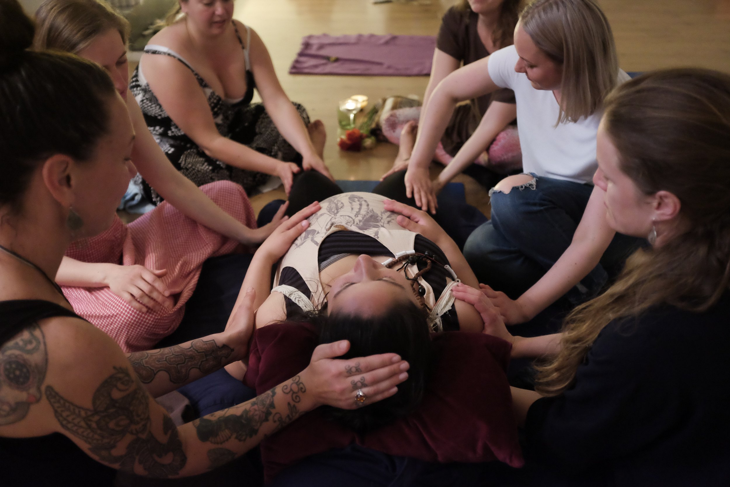 A group of women and a girl sitting on the floor, gathered around a person lying down, some with hands on the person, in a calming and spiritual setting.