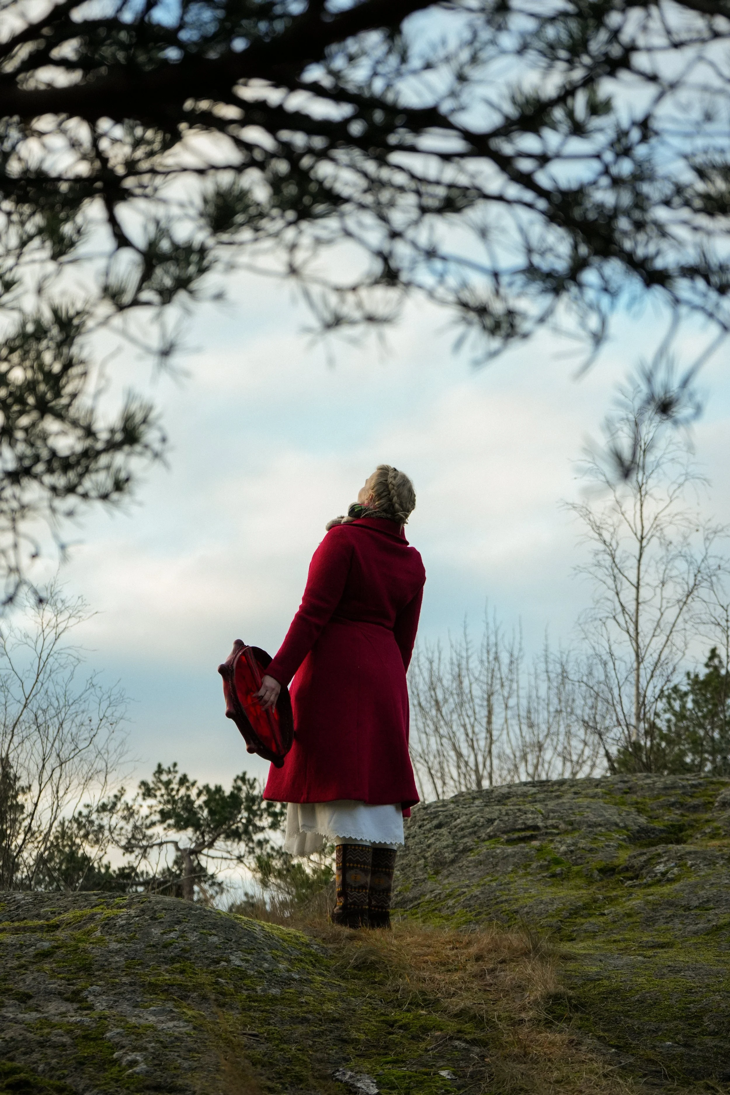 A woman dressed in vintage clothing, including a red coat, white skirt, patterned tights, and holding a red handbag, standing on a mossy rocky outcrop outdoors, looking up at the cloudy sky with bare trees and branches framing the scene.