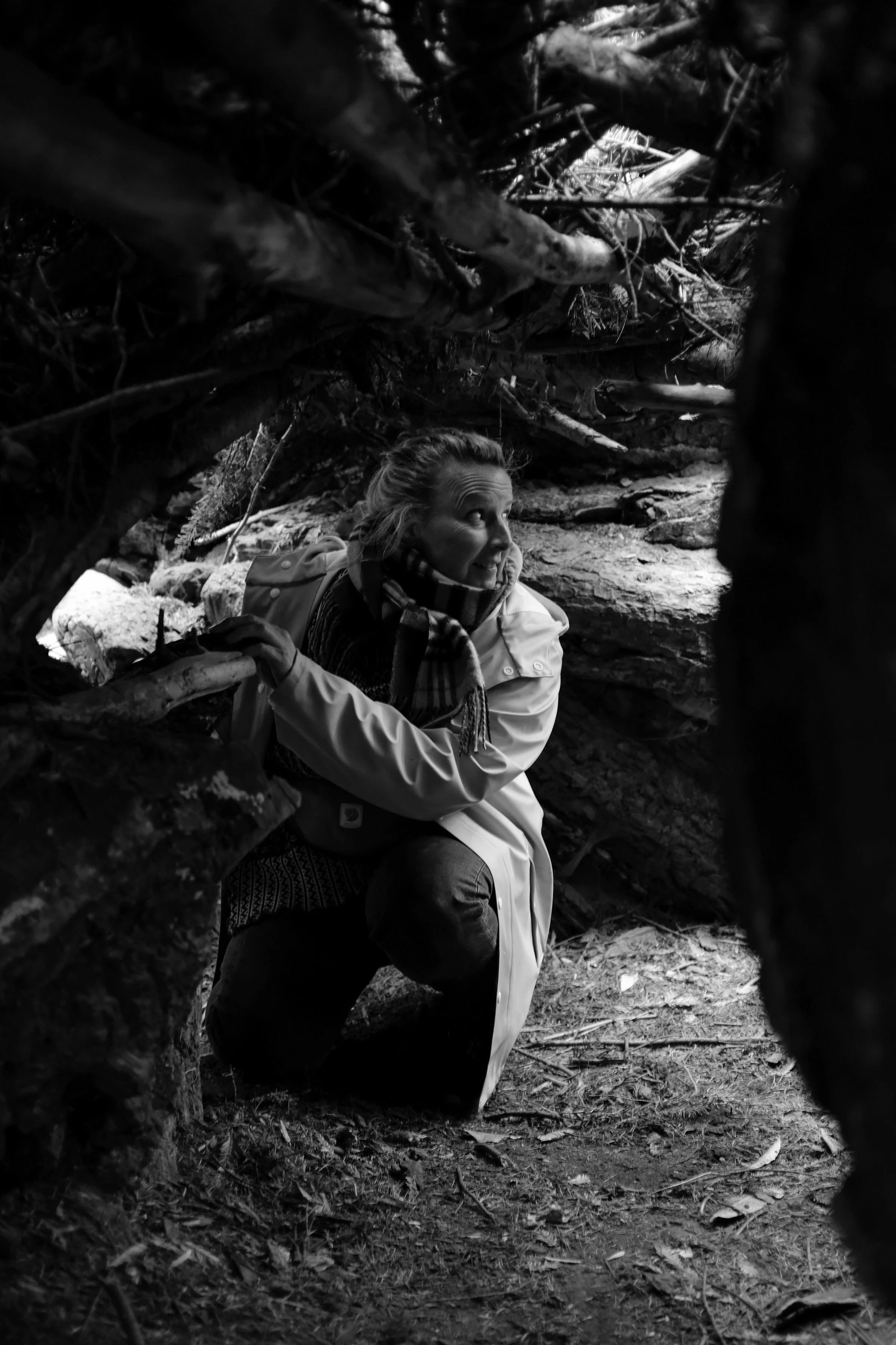 A woman in outdoor clothing and scarf, crouching among rocks and fallen branches in a forest, looking up and smiling.