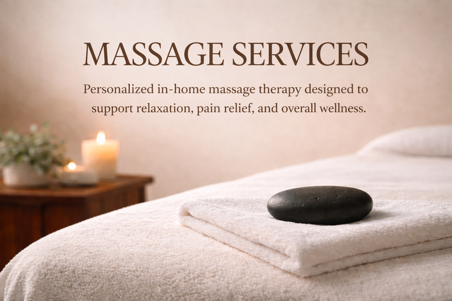 A massage table with a rolled-up towel and a smooth black stone, in a spa room with candles and a plant in the background.