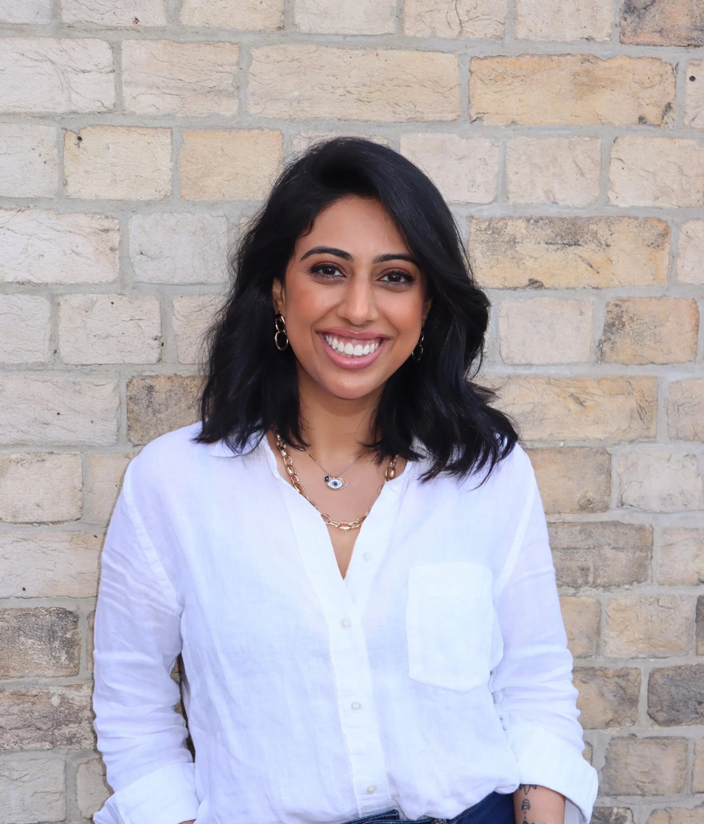 Smiling woman with dark hair wearing a white button-up shirt and jewelry standing in front of a brick wall.