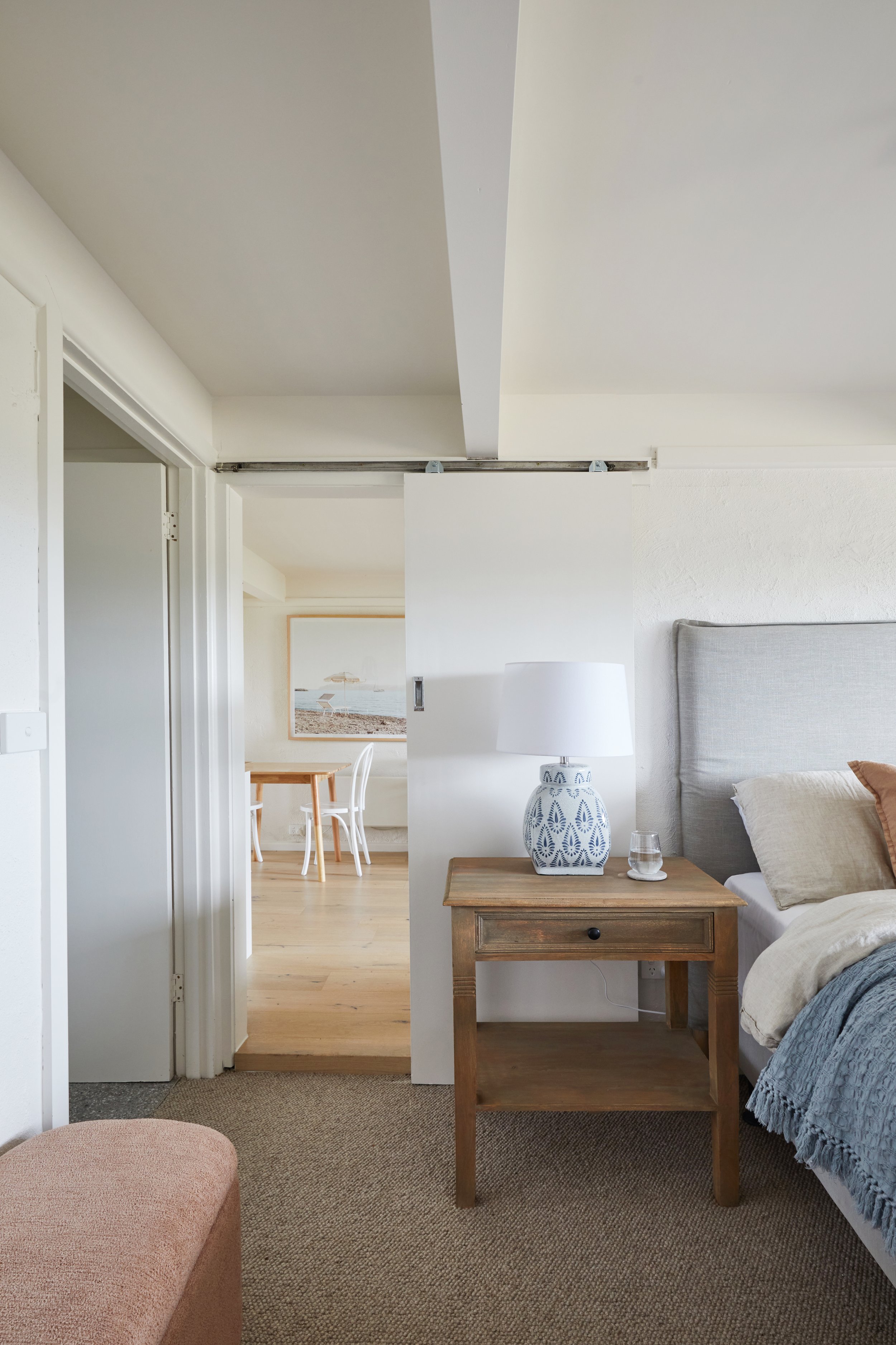 A bedroom with a wooden nightstand, a white lamp, a glass of water, and a bed with beige and blue bedding. There is a decorative box or container on the nightstand. In the background, a doorway leads to a dining area with white chairs and a framed picture of a beach scene on the wall.