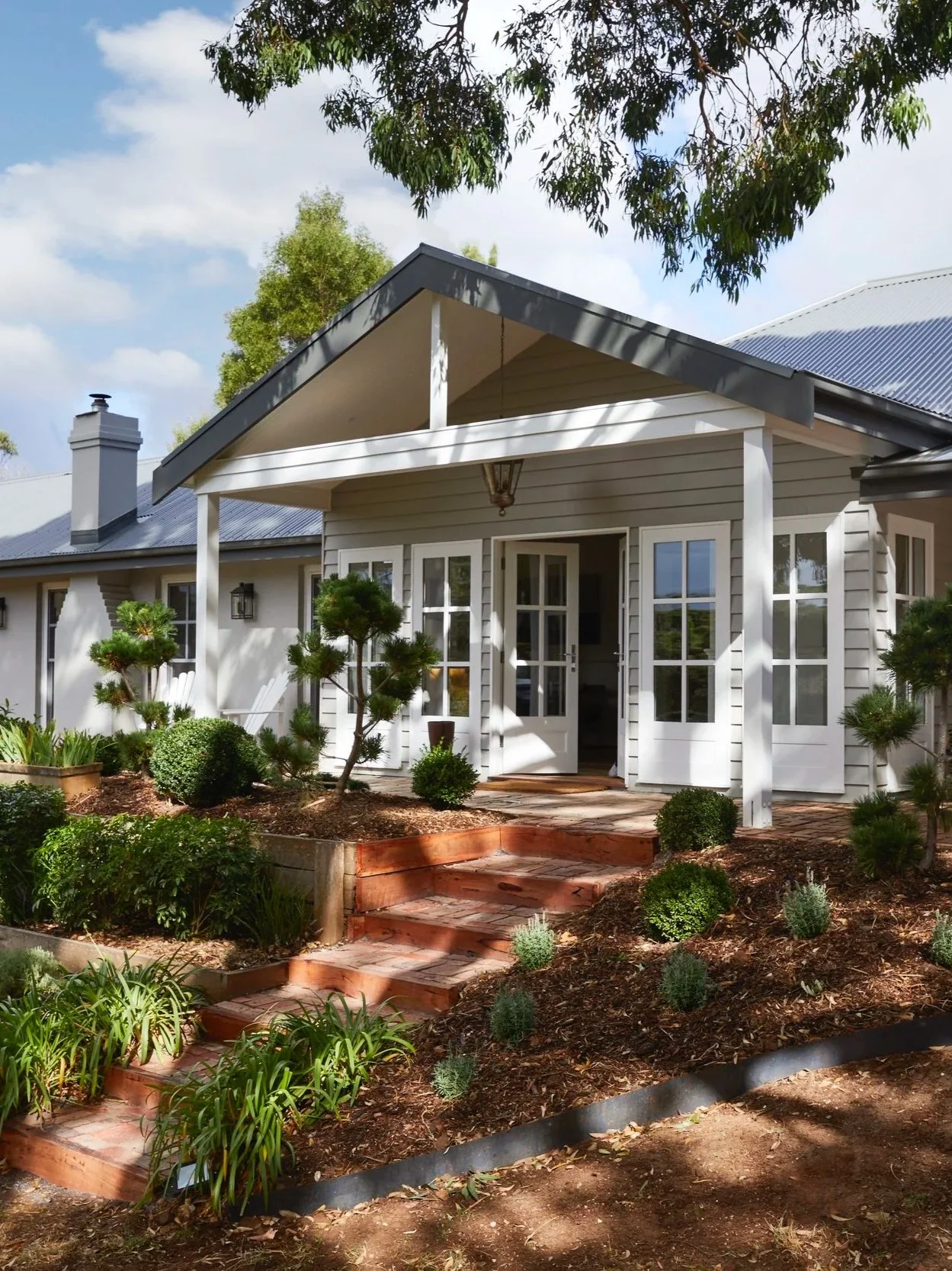 A white house with a covered porch, a manicured front yard with small bushes, trees, and a brick pathway leading to the entrance, under a blue sky with clouds.