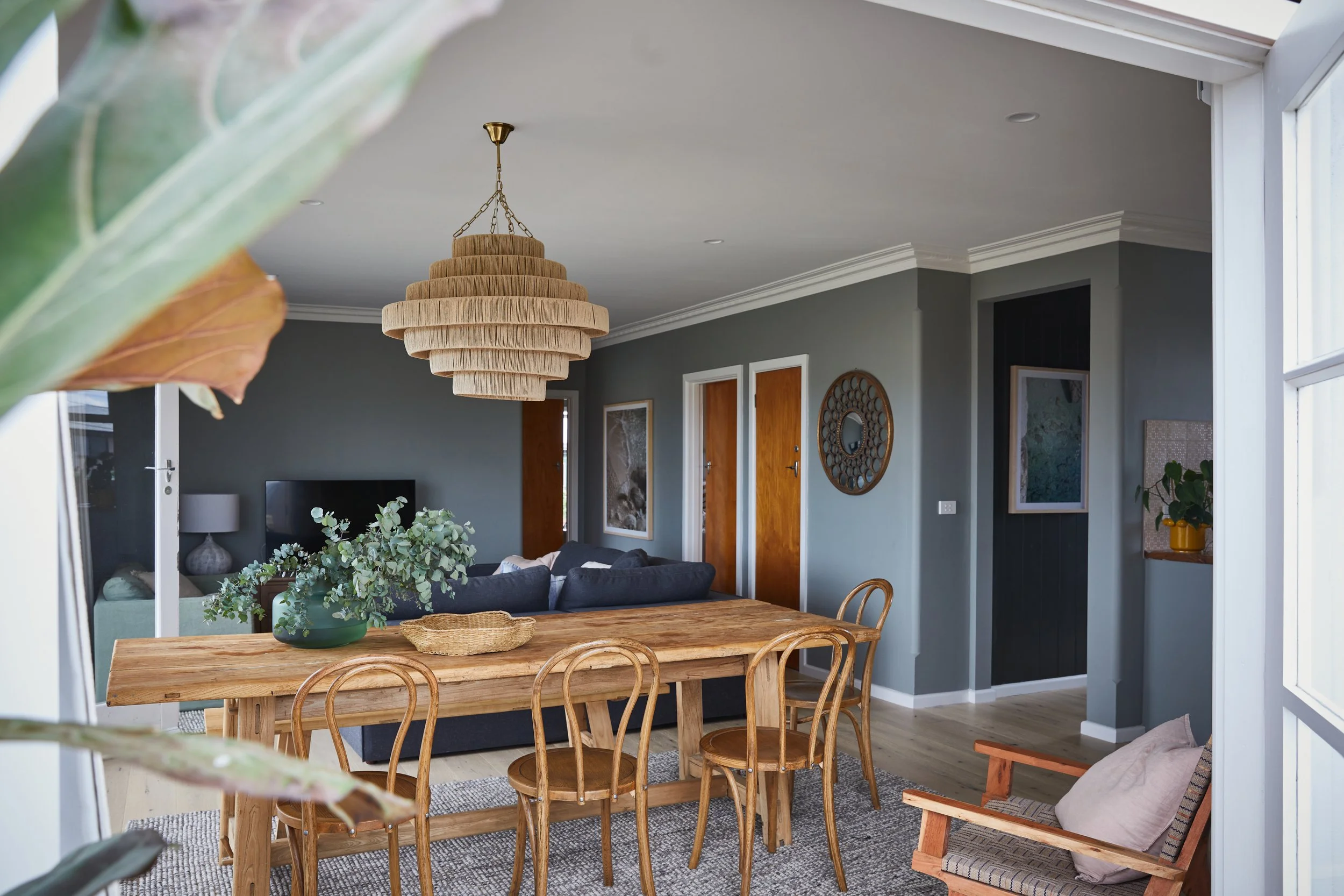 A living and dining room with a wooden dining table, matching wooden chairs, a gray sofa, artwork on the walls, a wicker pendant light, and leafy plants near a large window.