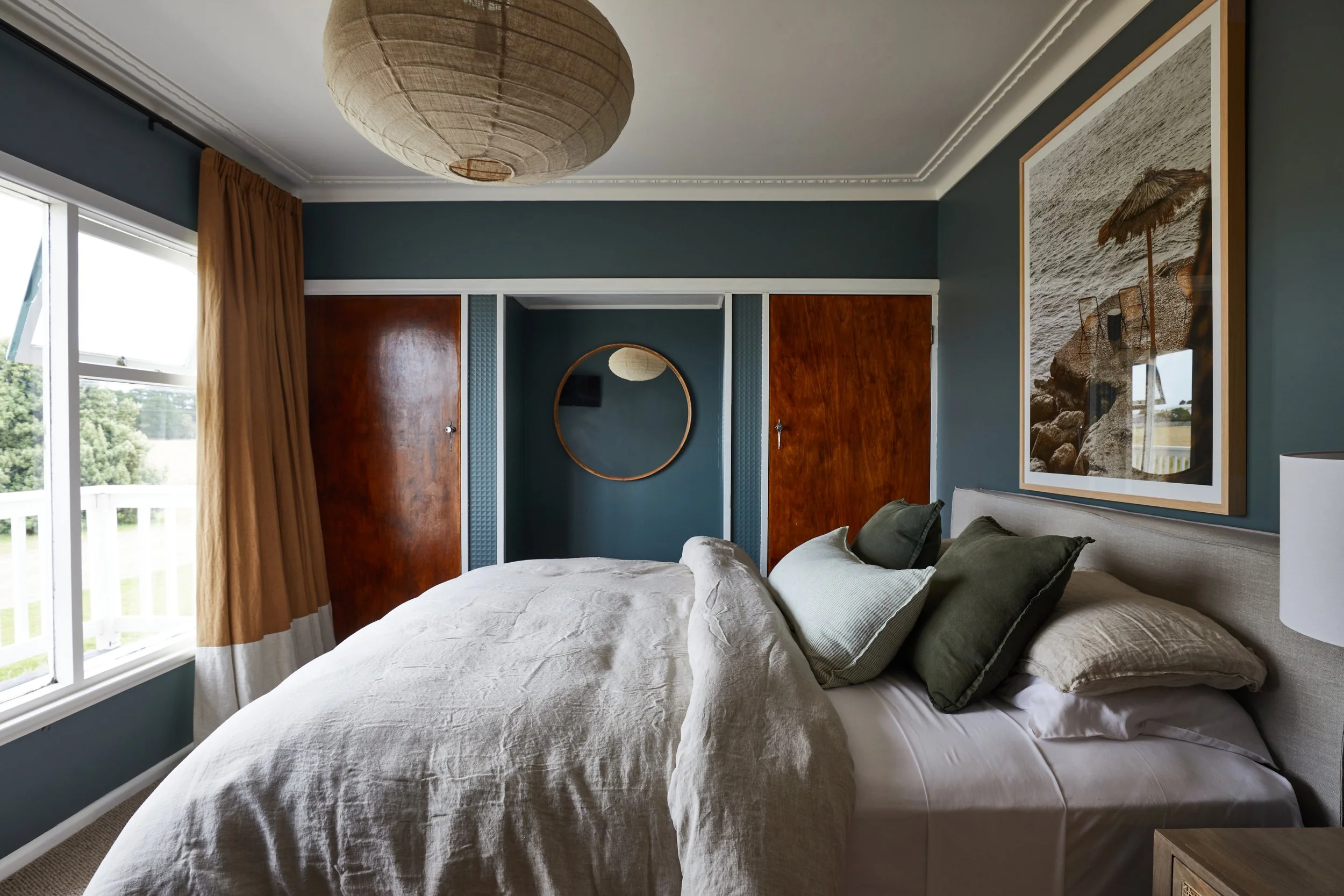 A bedroom with a large bed covered in white bedding and multiple pillows, a round mirror above the bed, a large framed beach photo on the wall, a window with beige curtains, a wooden closet door, and a paper lantern ceiling light.