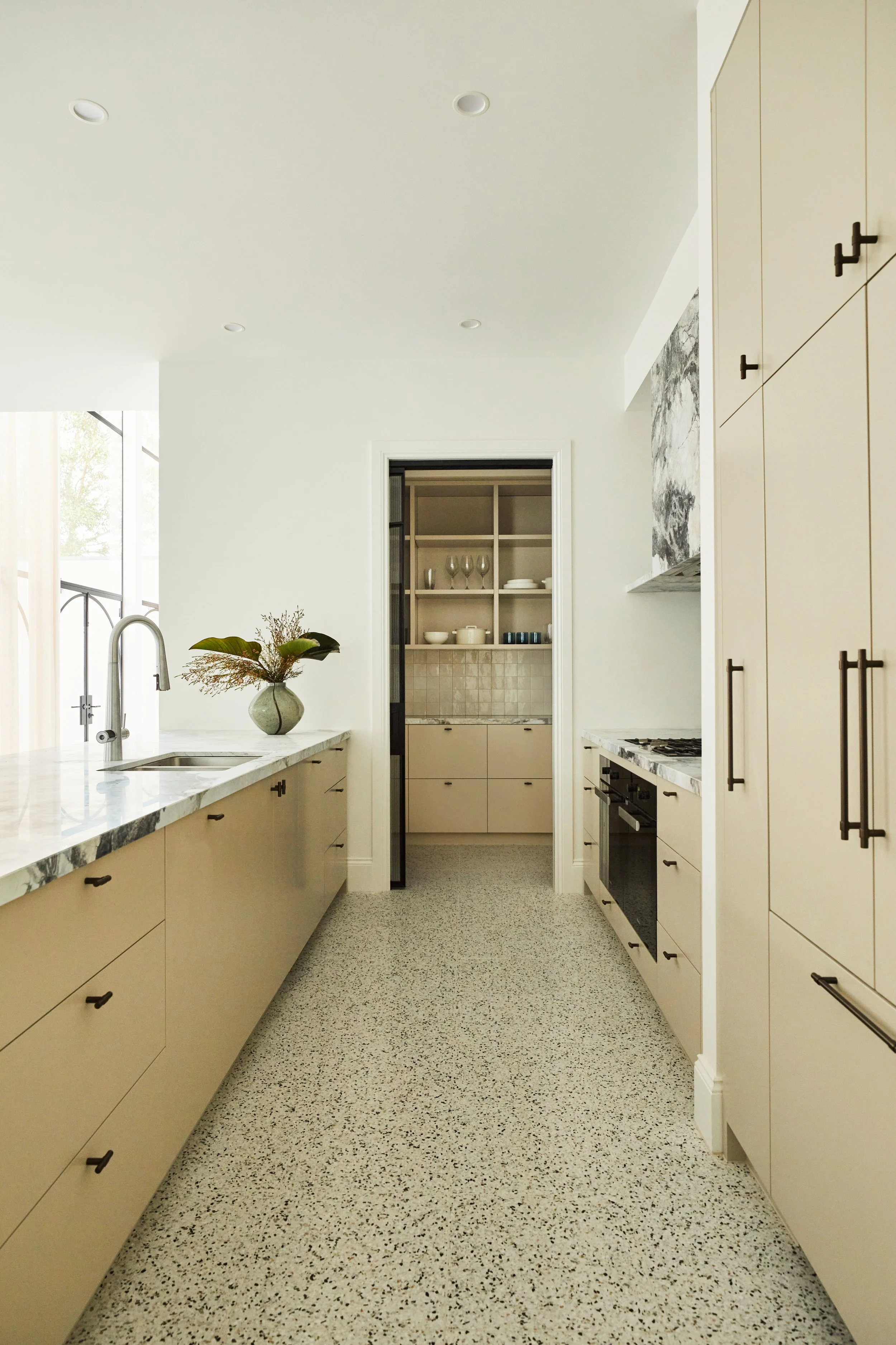 A modern kitchen with beige cabinets, black handles, marble countertops, and a vase with flowers on the counter. There is a doorway at the end leading to another room with shelves and dishes visible. The floor is speckled terrazzo, and the ceiling has recessed lighting.