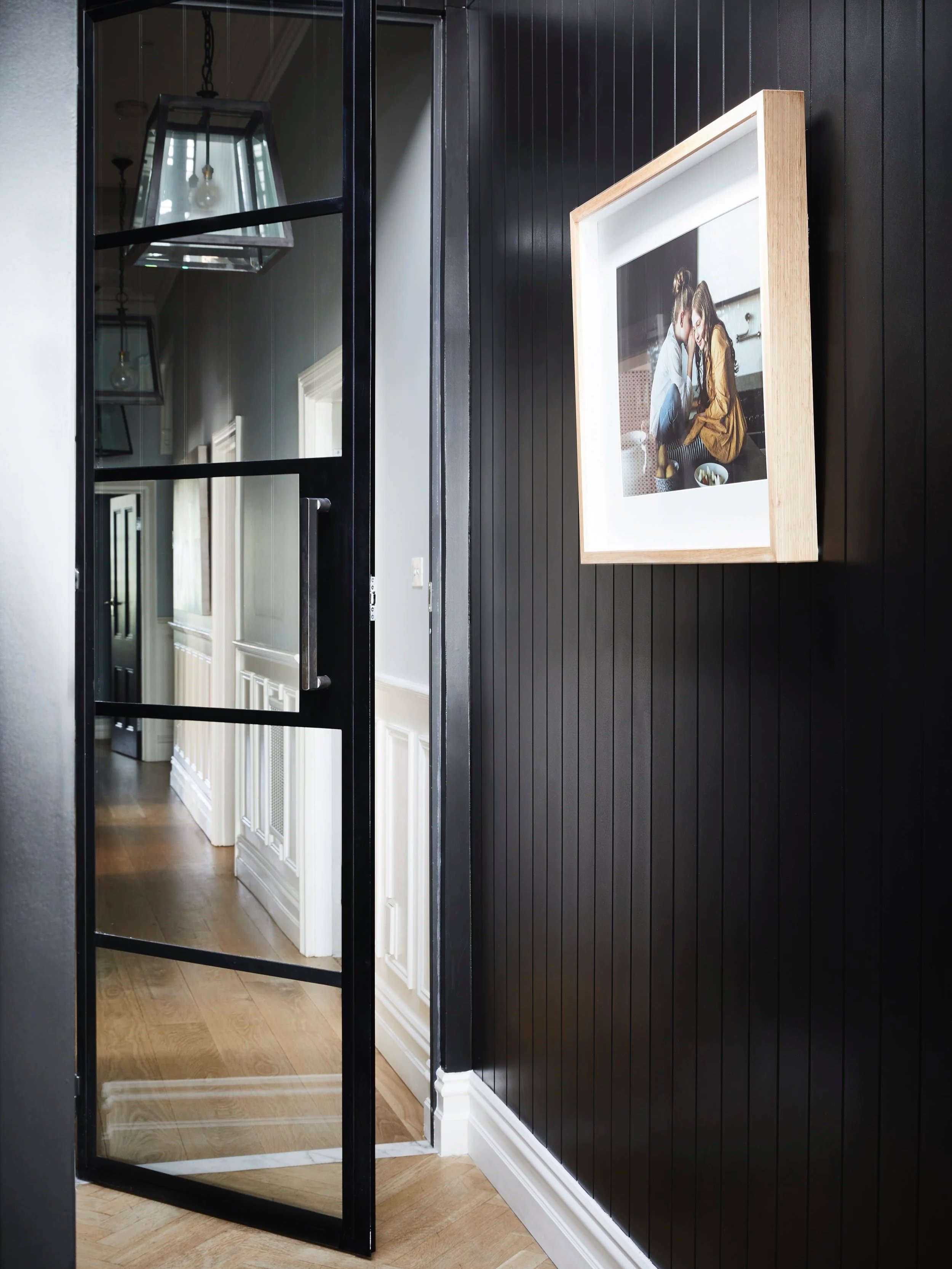 A black-framed glass door with a handle, opening into a room with white cabinets and hardwood flooring, with a black wall and framed artwork on the wall.