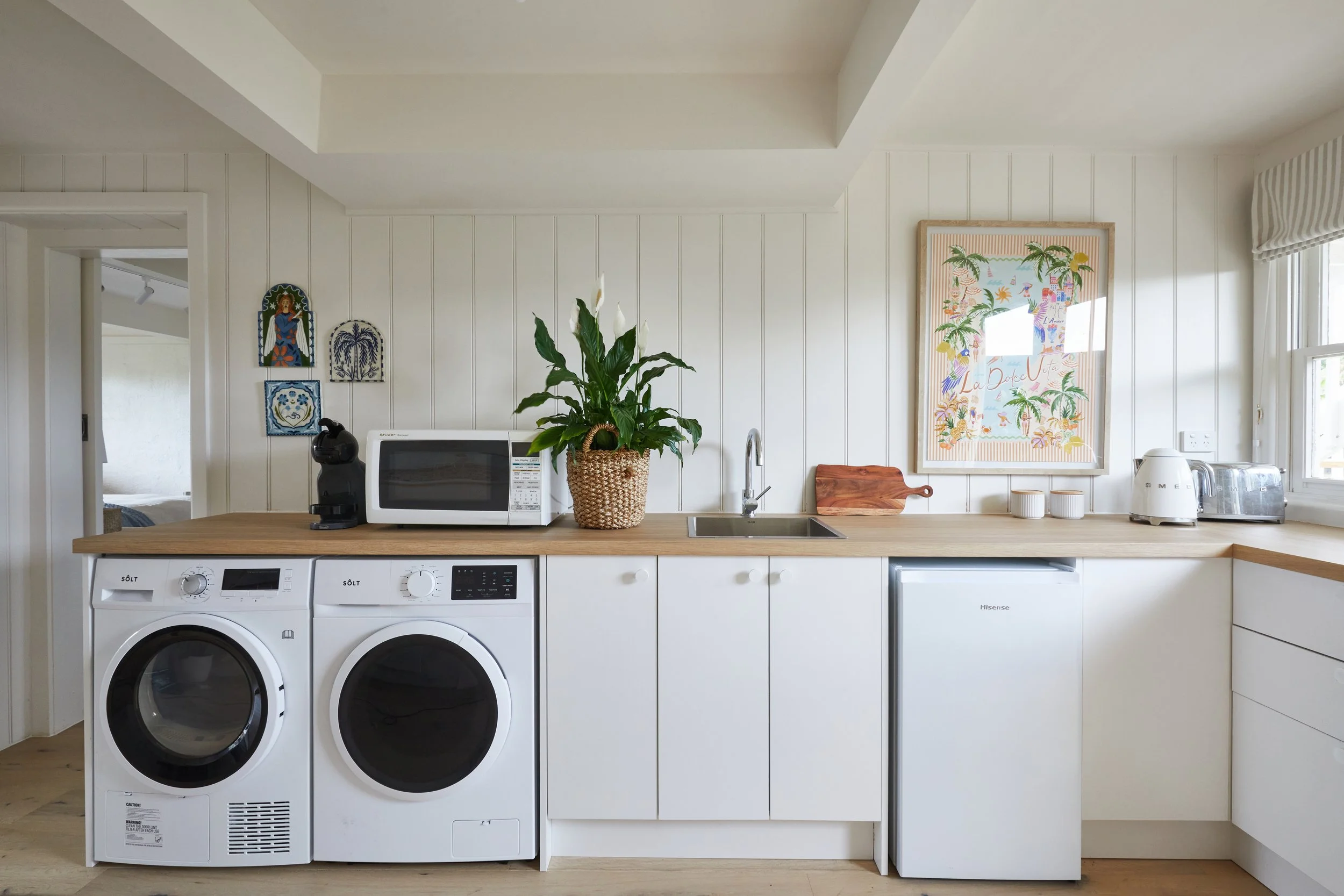 A kitchen with white walls and wooden countertop. On the countertop, there are a microwave, a potted plant, a cutting board, two small containers, a toaster, and a coffee maker. Below the counter, there are two washing machines. There are framed artworks on the wall, and a window with striped curtains.