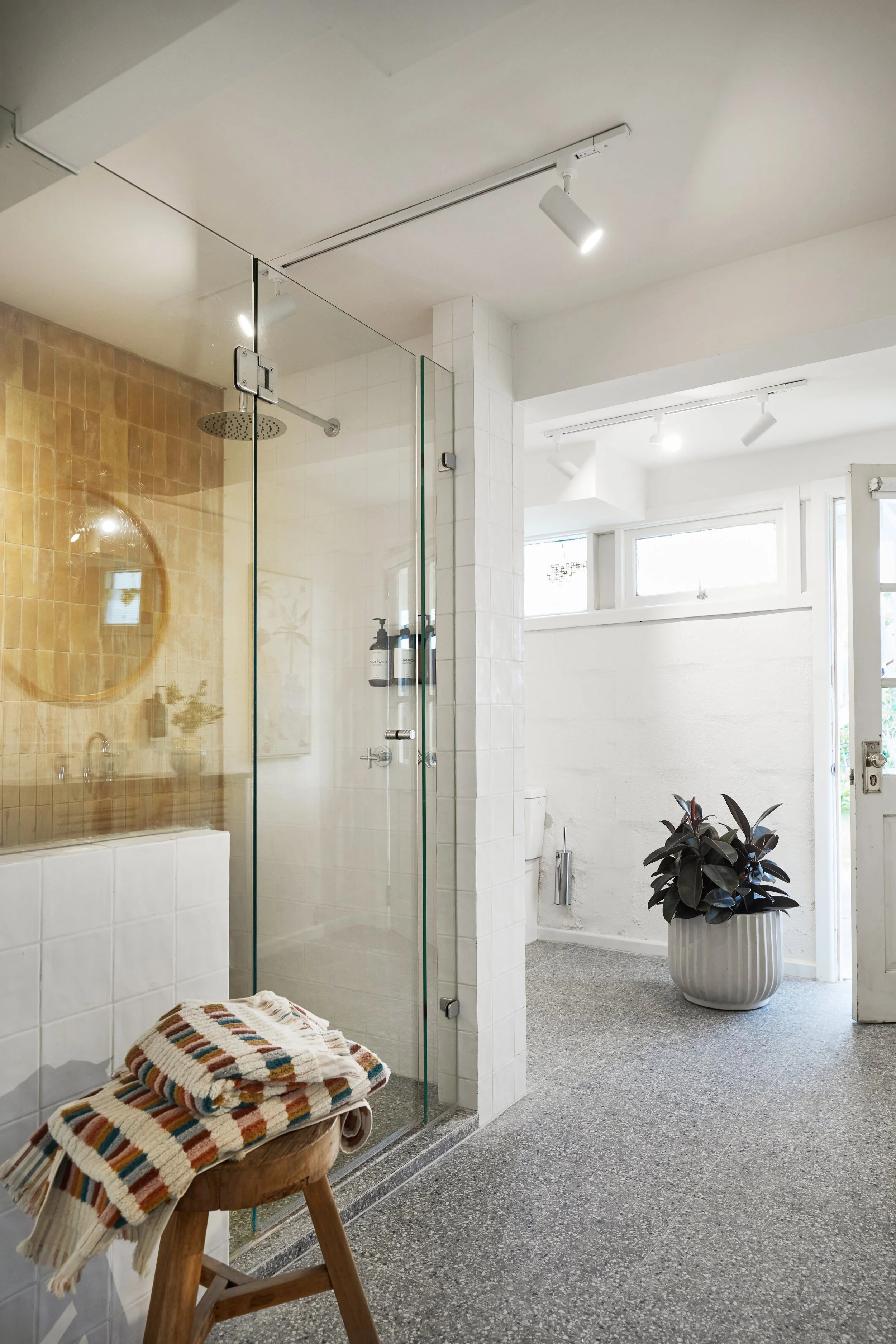 A modern bathroom with a glass shower enclosure, a potted plant in a striped planter, natural light from large windows, and a cozy stool with a striped blanket.