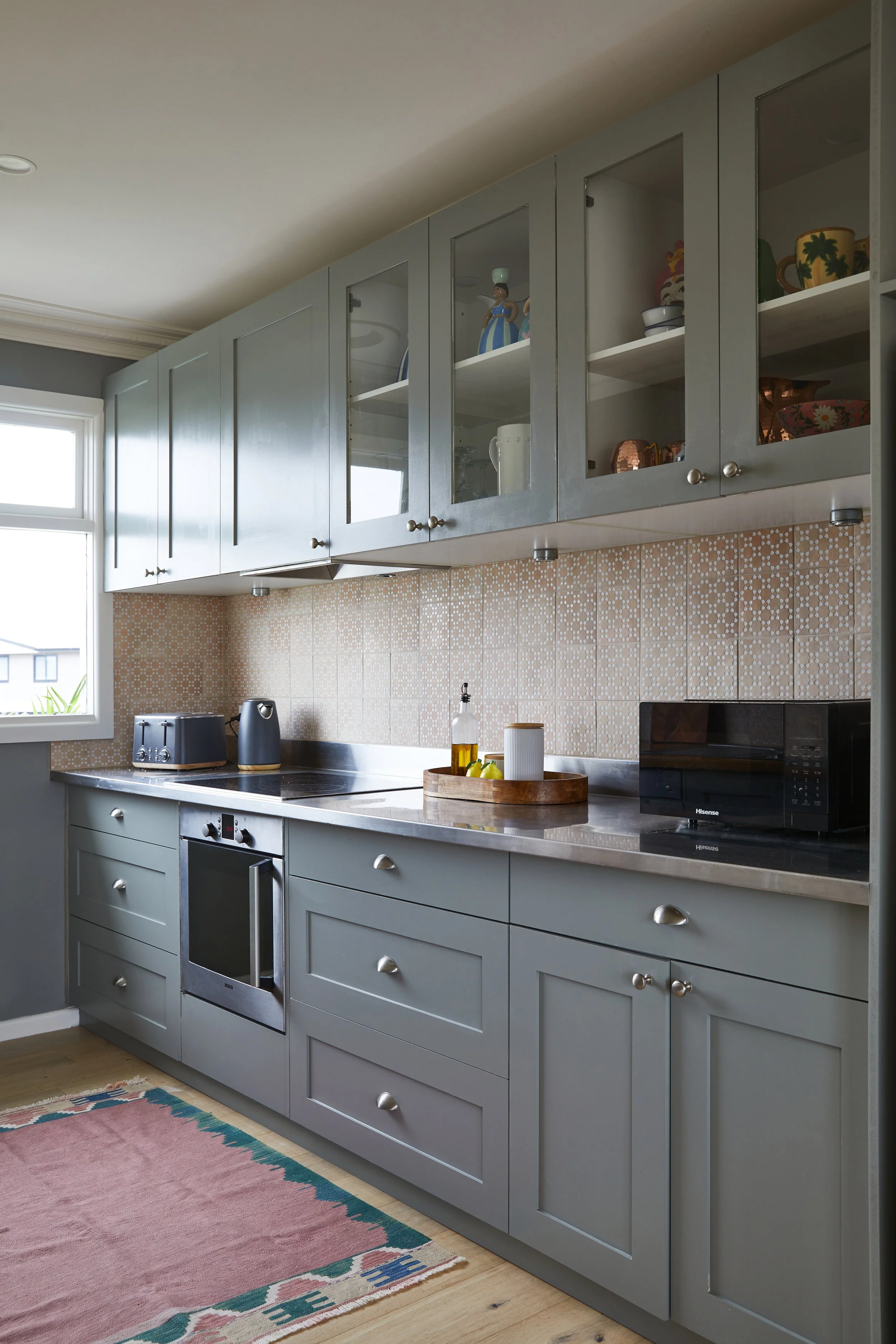 Kitchen with gray cabinets, some with glass doors displaying decorative items, a stainless steel oven, black appliances, a wooden tray with oil and lemons on the counter, and a colorful rug on a light wood floor.