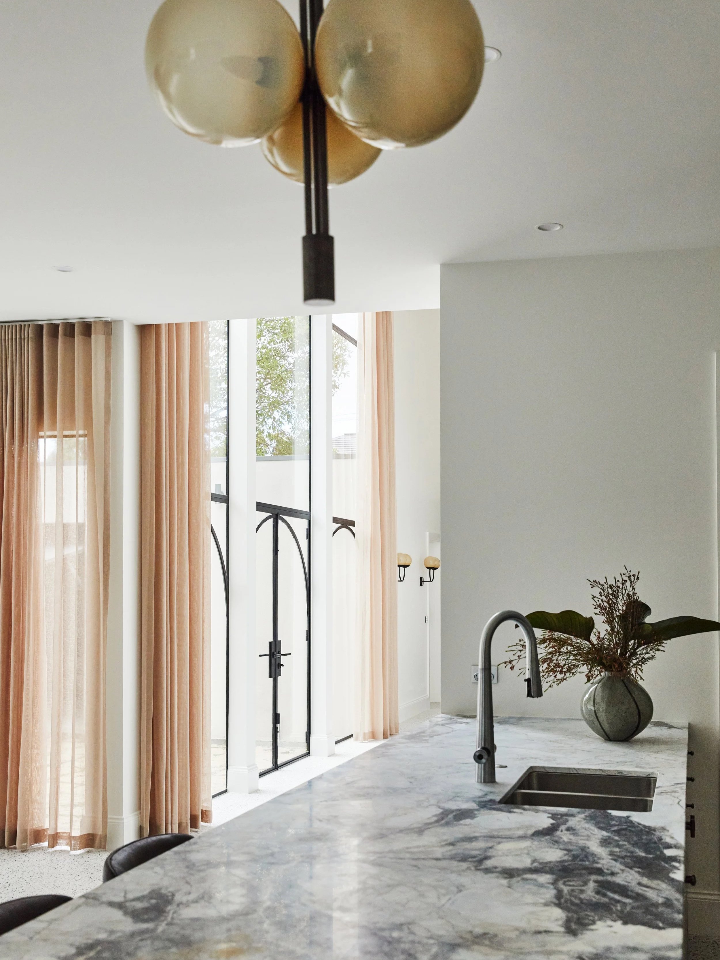 Modern kitchen with marble countertop, stainless steel sink, black faucet, large windows with sheer peach curtains, glass doors, and a decorative vase with dried flowers.