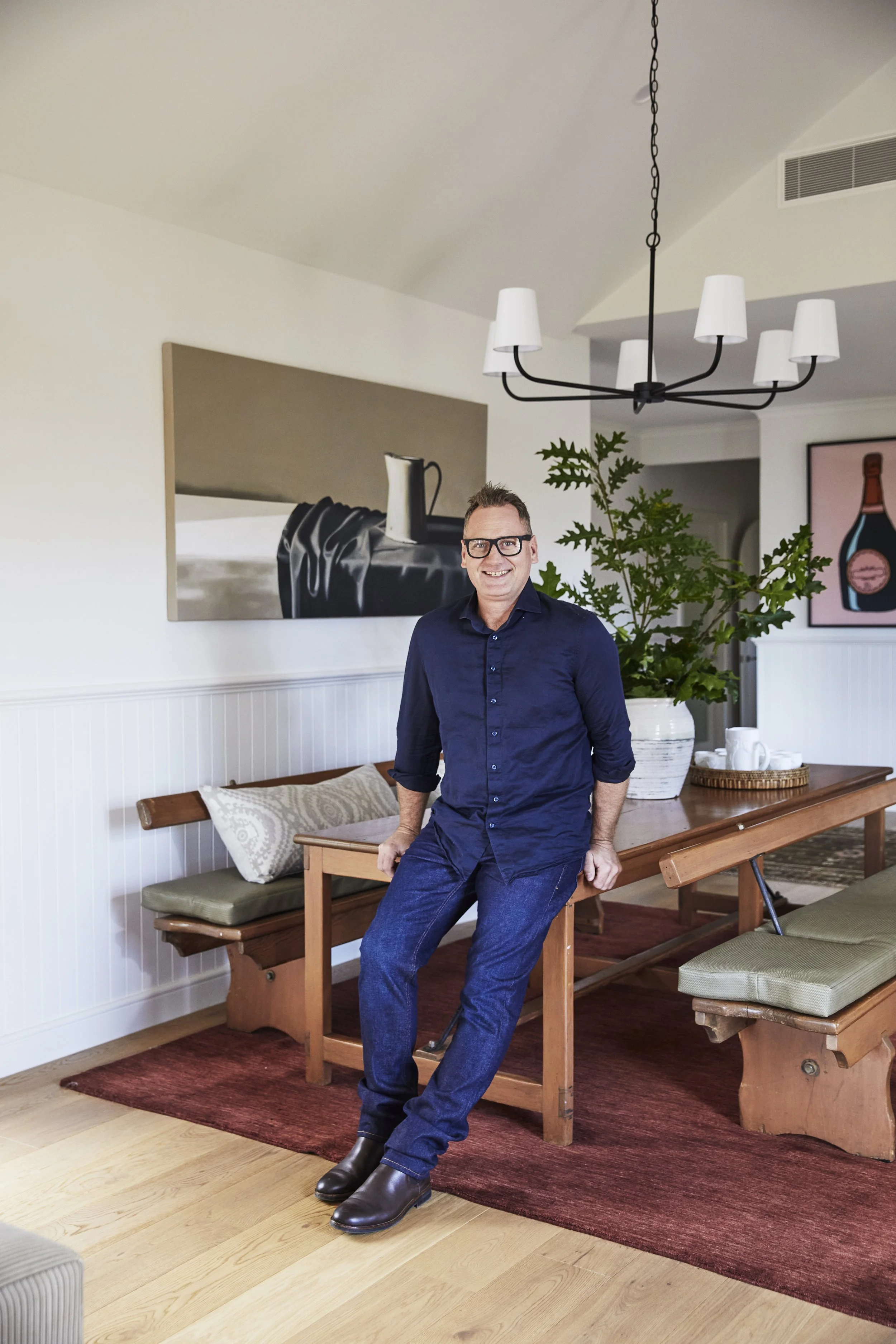 Luke Shannon with glasses wearing a dark blue shirt and jeans sitting on the edge of a wooden dining table in a living room.