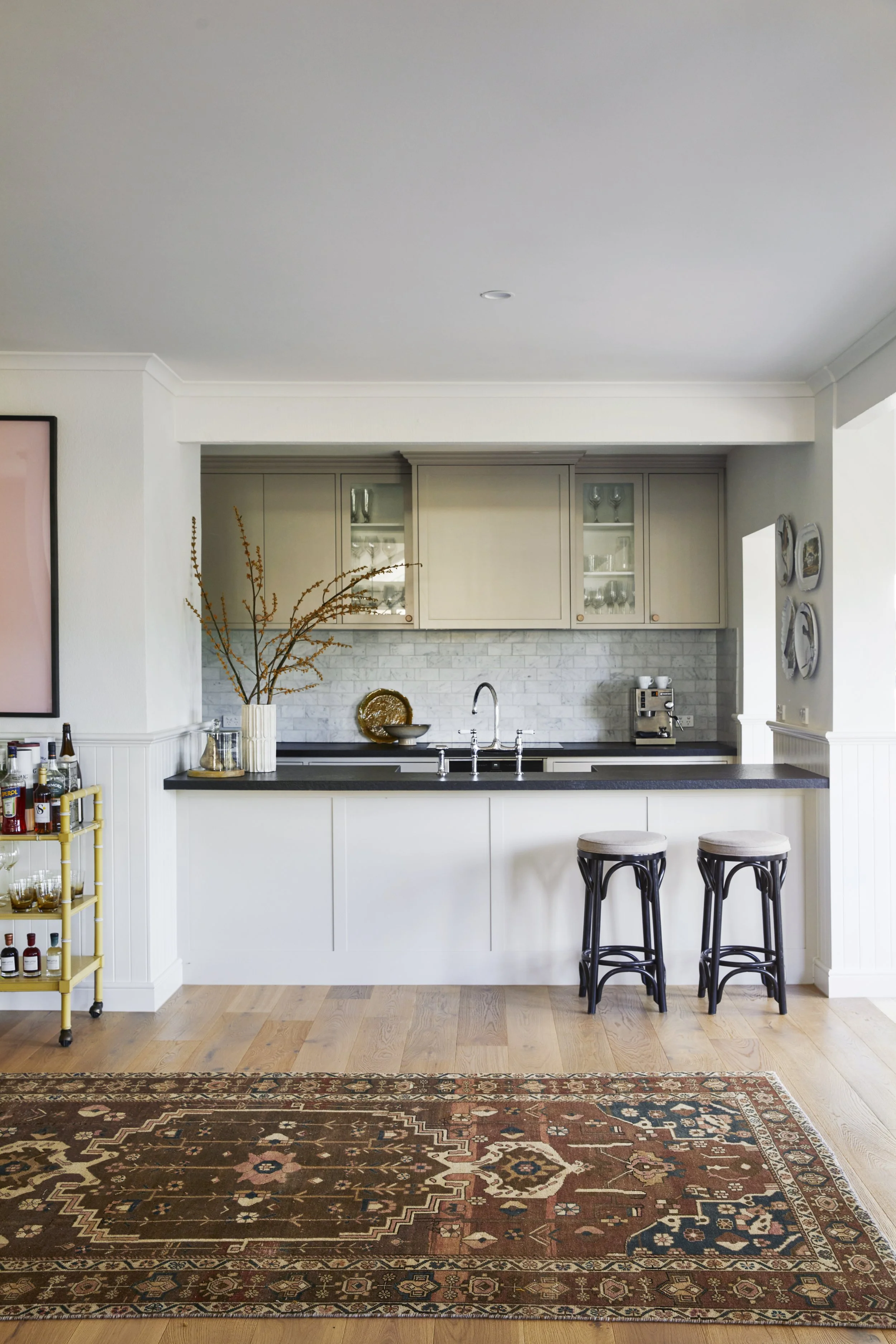The image shows a modern kitchen with a black countertop, light-colored cabinets, glass-front upper cabinets, and a backsplash of white subway tiles. There are two leather and metal stools at the counter, a decorative vase with dried branches, a round decorative tray, a stack of white bowls, a coffee machine, and a colorful oriental rug on the wooden floor.
