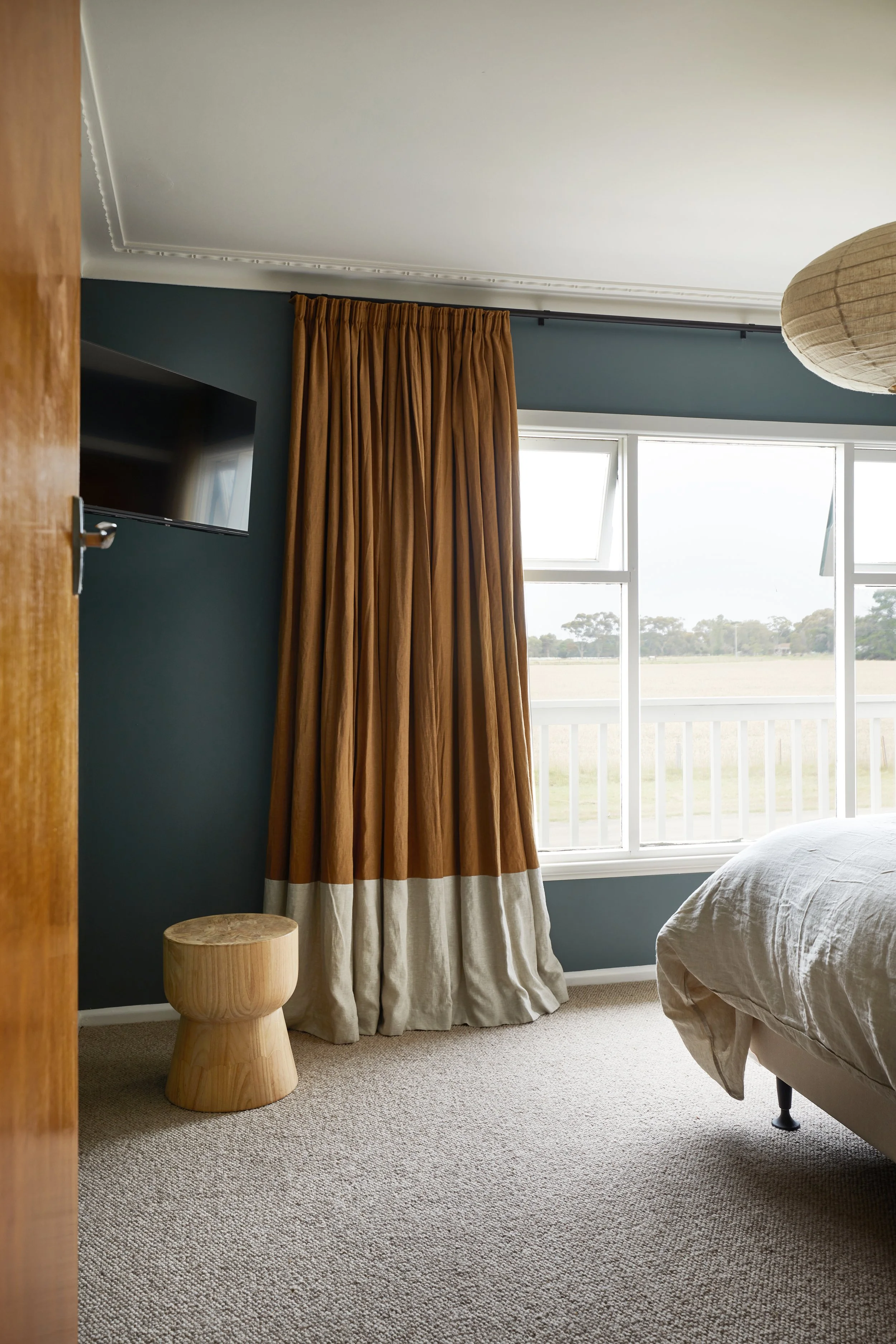 A bedroom with beige carpet, a partially visible bed with white bedding, dark green walls, a window with open screens showing a grassy field and trees outside, gold curtains, a wooden stool, and a wall-mounted television.
