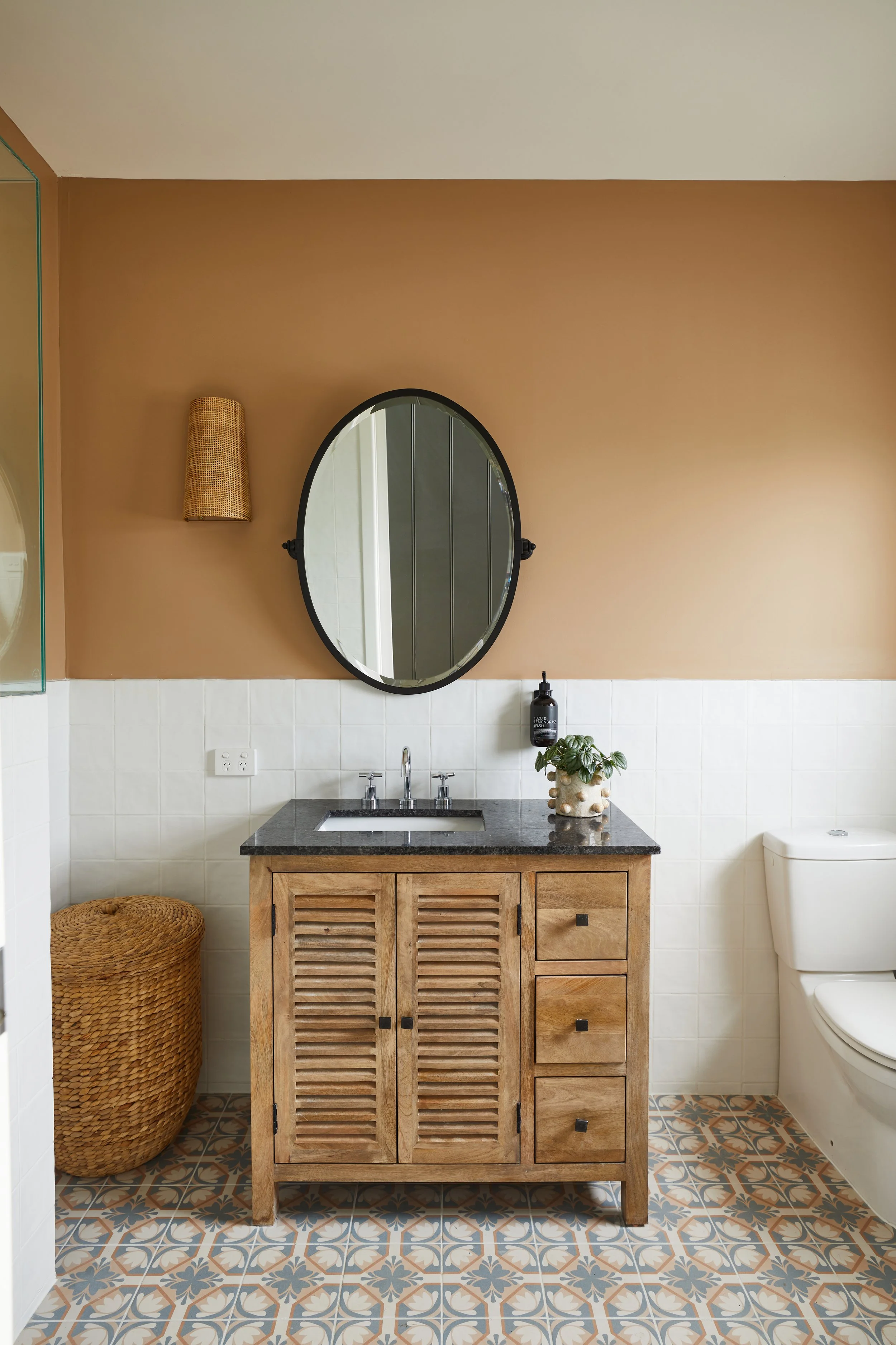 A bathroom with a wooden cabinet under the sink, a mirror above the sink, and a basket on the floor. The wall is painted brown with a light fixture and a soap dispenser, and a toilet is visible on the right. The floor has patterned tiles.