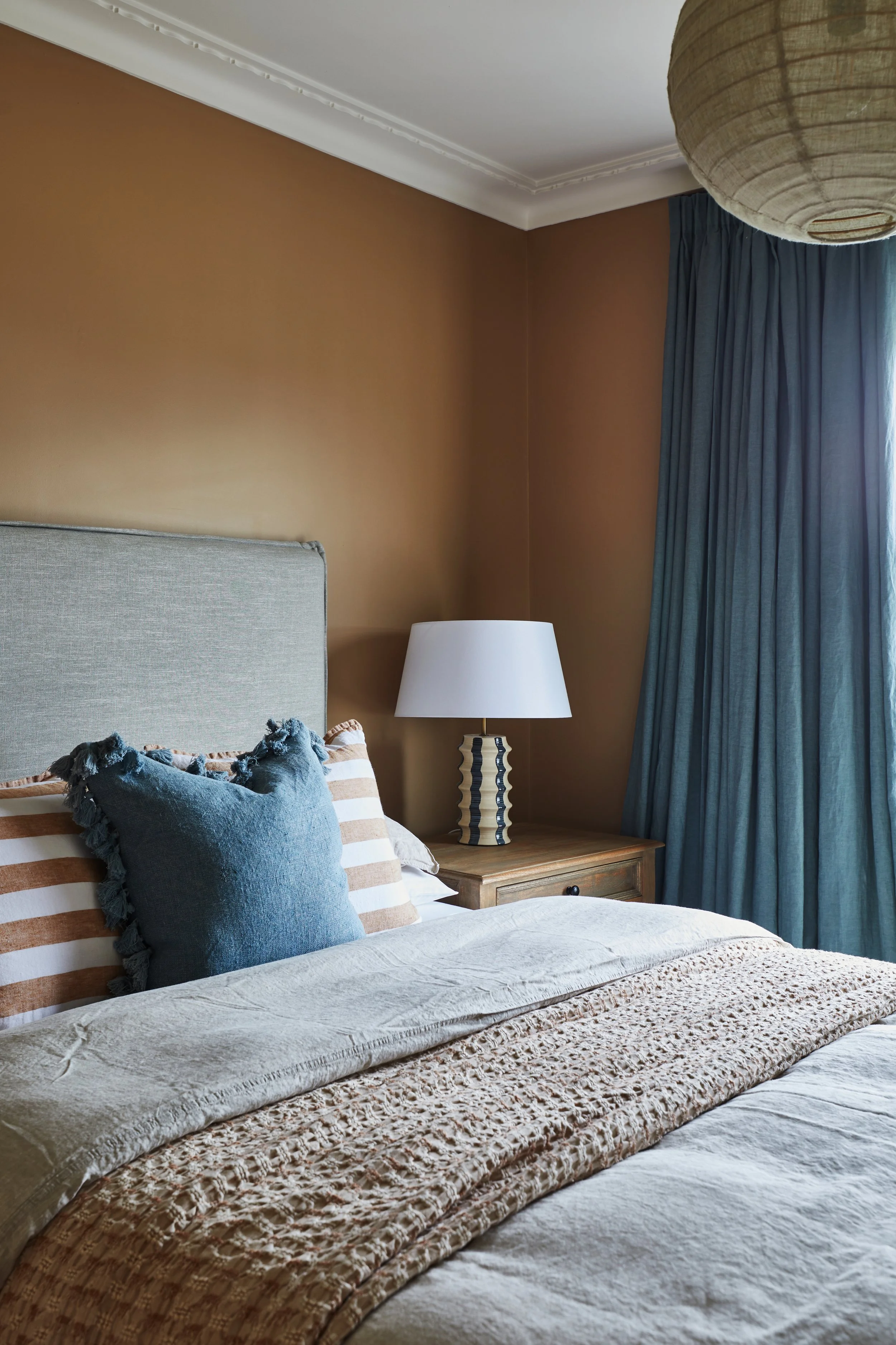 A neatly made bed with decorative pillows, featuring a beige crocheted blanket on top, in a bedroom with an orange accent wall, blue curtains, a wooden nightstand, and a white lampshade.